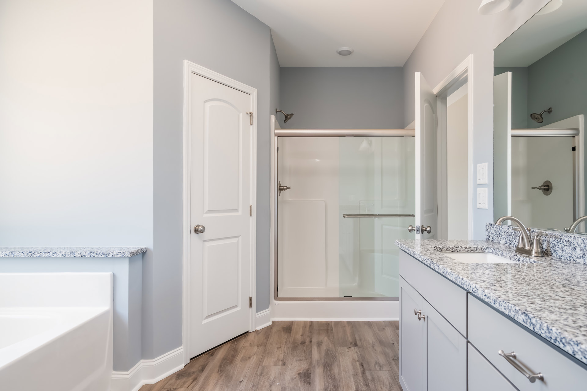 Bathroom with glass shower enclosure, white bathtub, wall-mounted sink beneath a large mirror, white door with silver knob, light tile flooring and walls, chrome shower head and