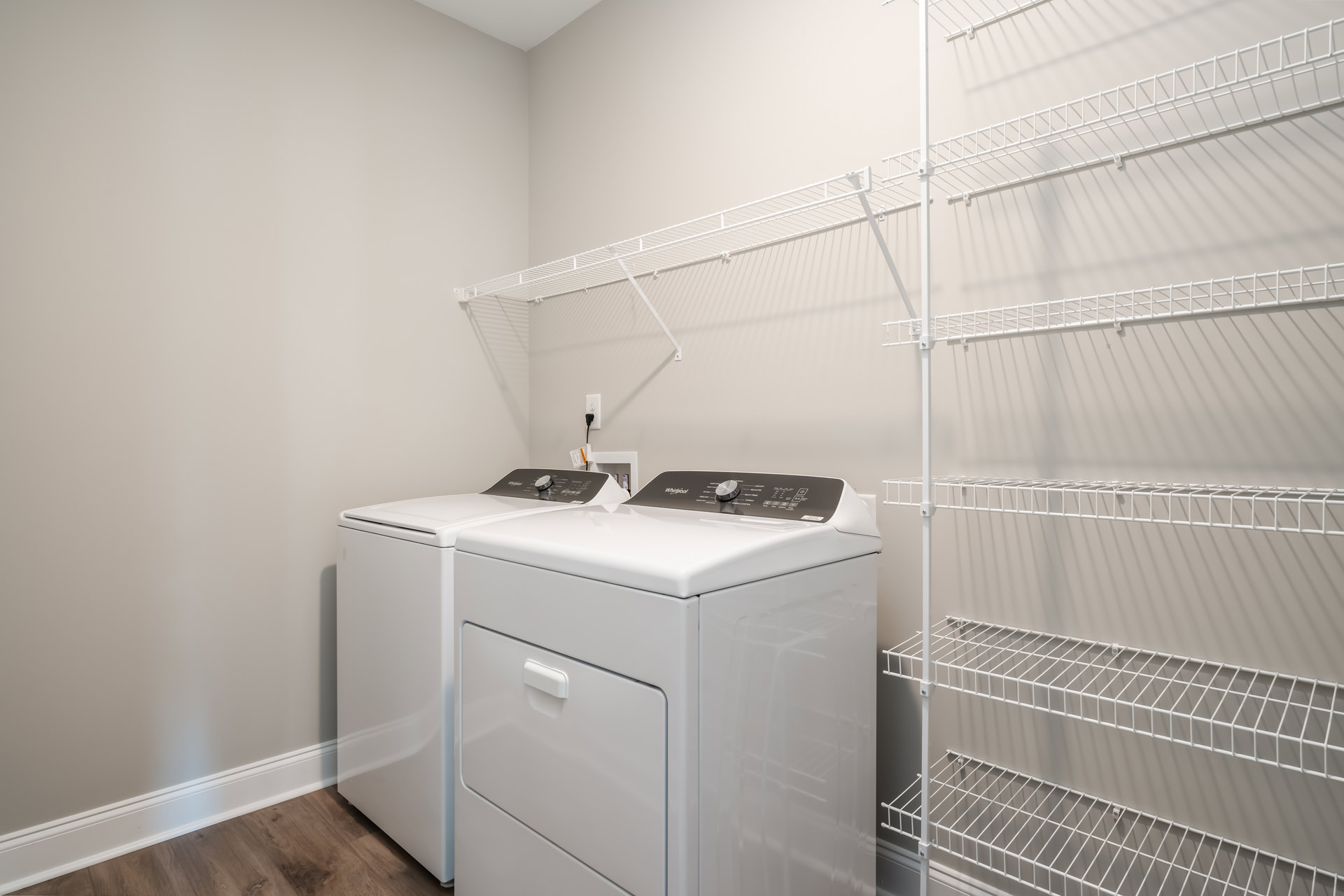Front-loading washing machine and dryer set beneath white wall-mounted shelves in a laundry room with light cabinetry and neutral walls