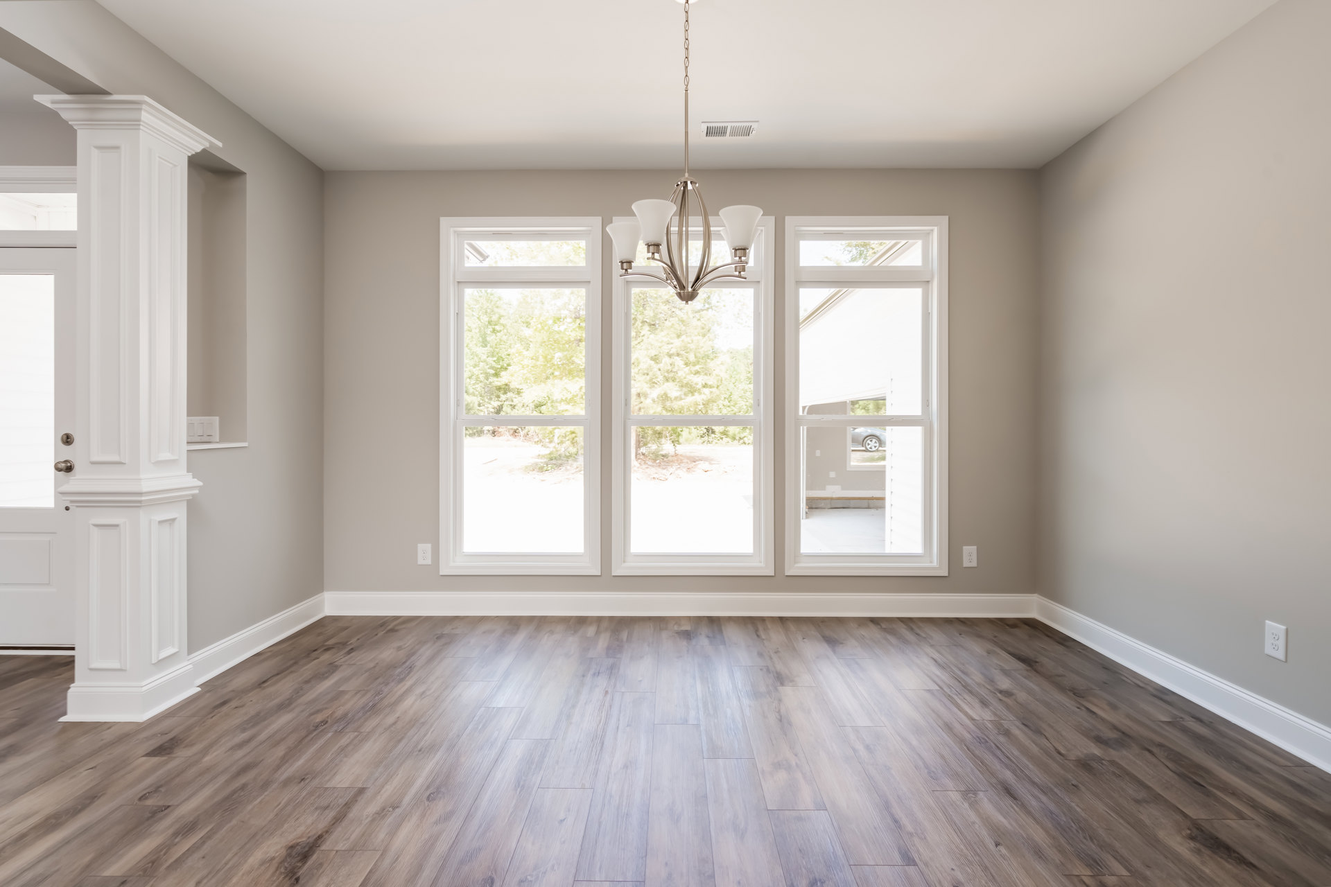 Chandelier hanging from white ceiling above hardwood floor with white trim, large windows letting in natural light, parked car visible outside