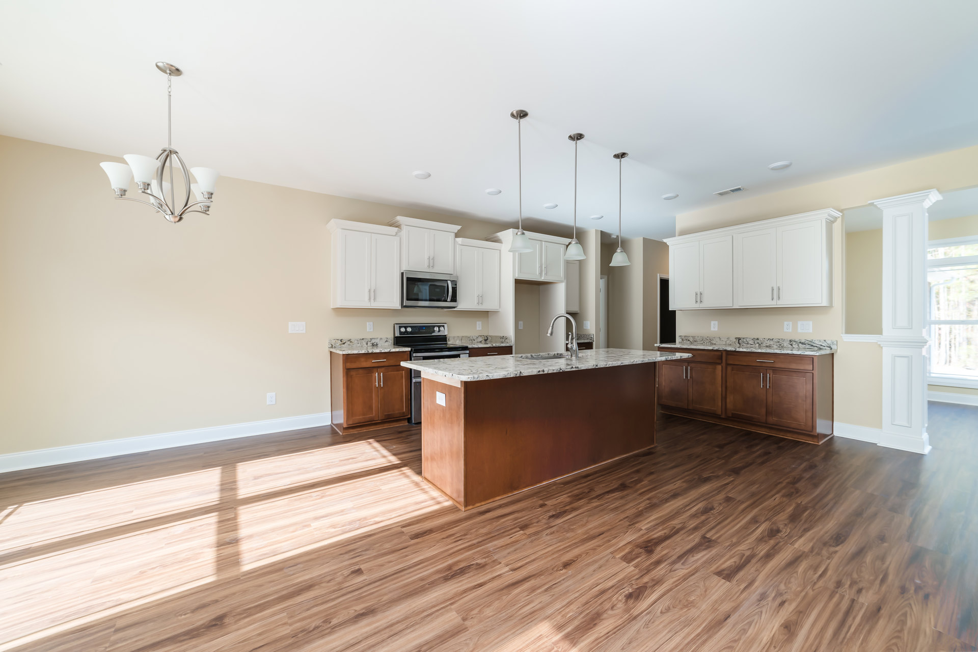 Spacious kitchen featuring a large central island with built-in sink, wood flooring illuminated by natural light, white cabinets with silver handles, microwave with white door, and