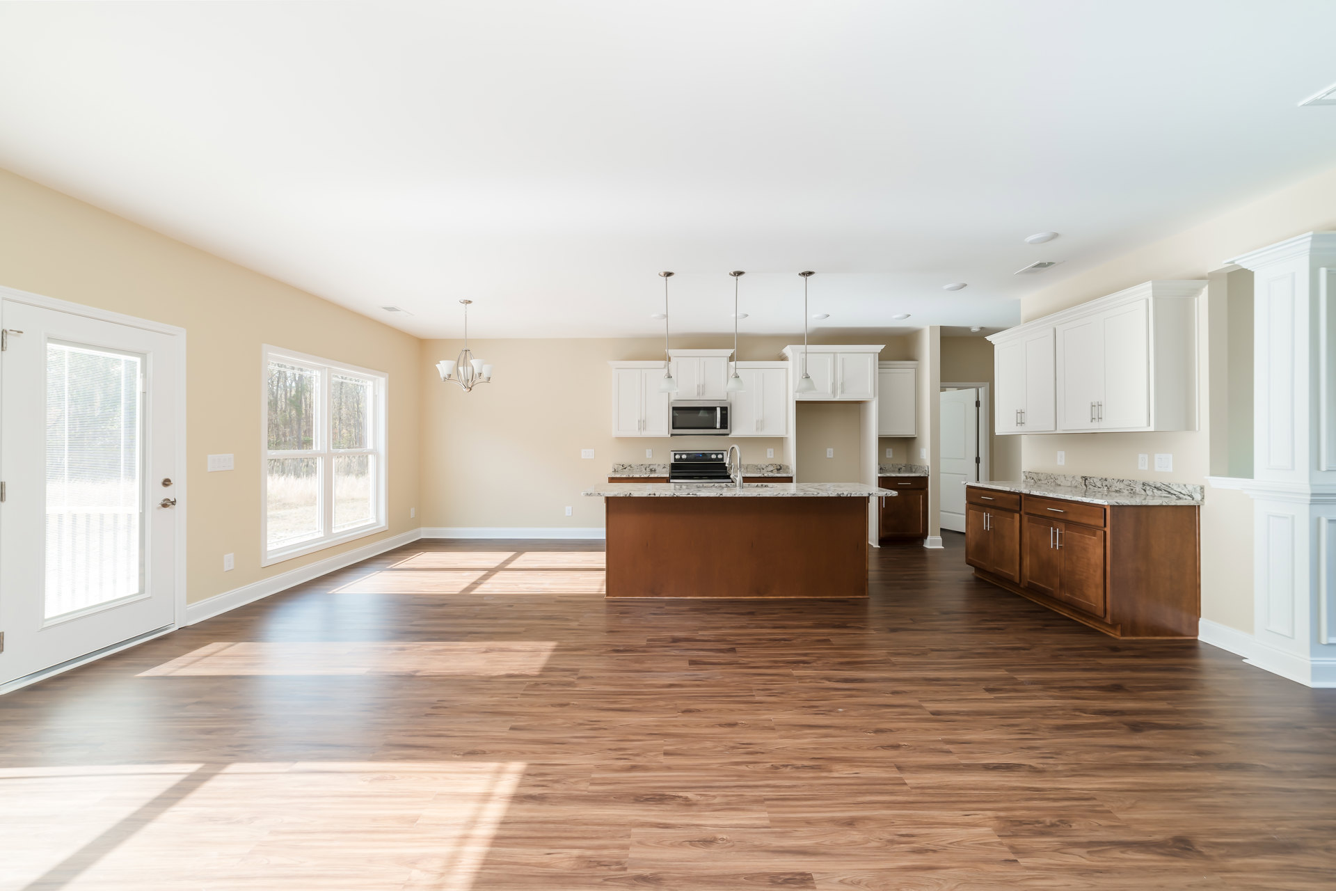 Spacious kitchen featuring wood flooring, a central island with stone countertop, white cabinetry, stainless steel oven, large window overlooking trees, and a white door with