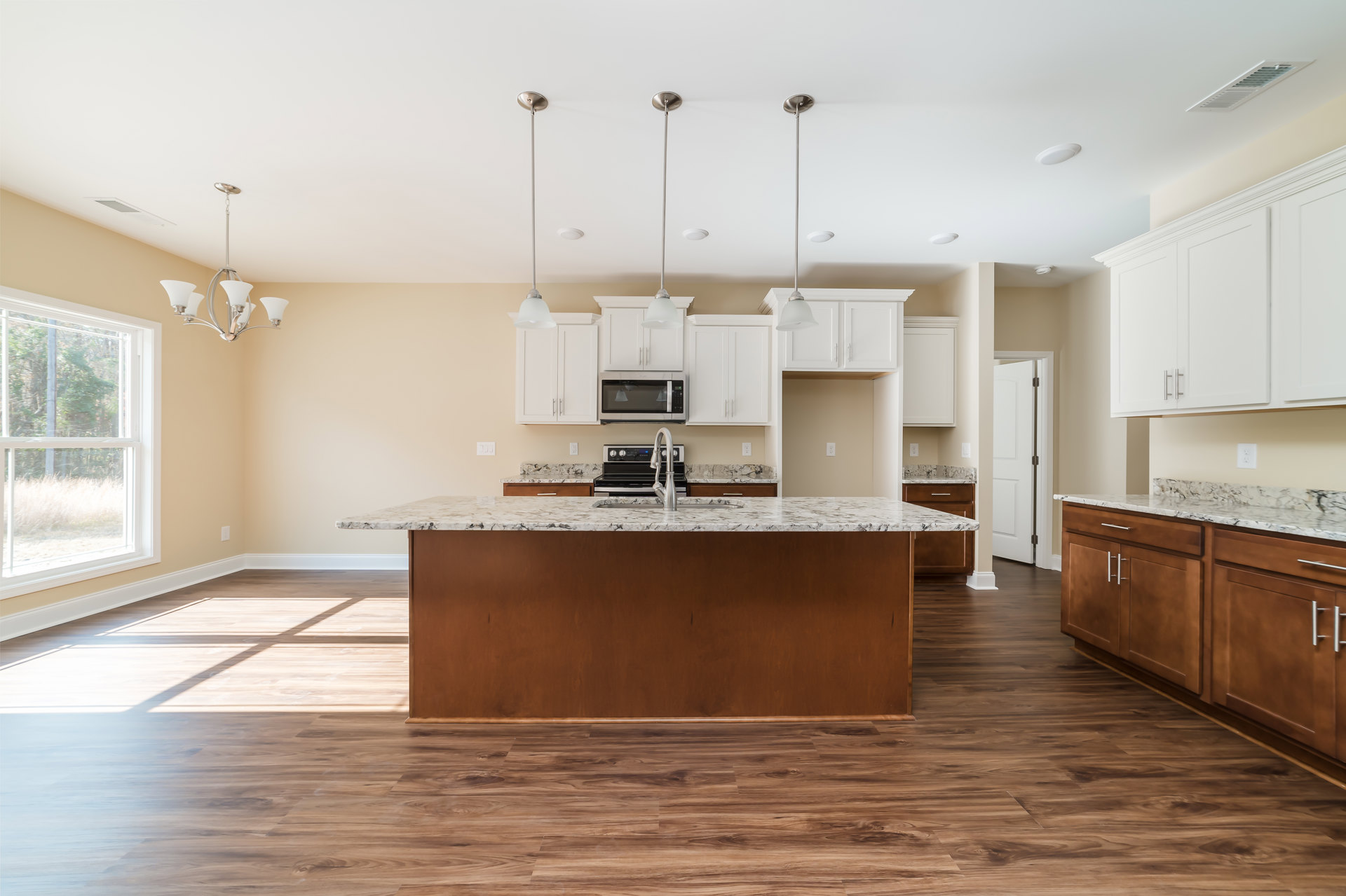 Spacious kitchen featuring a large central island with white cabinetry, stainless steel sink and faucet, glass door microwave, stove, and a window overlooking leafy trees; light