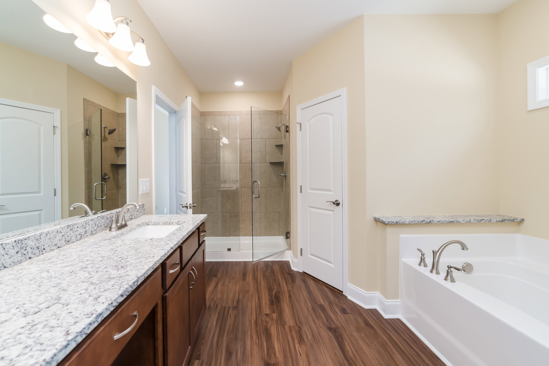 Bathroom featuring a frameless glass shower, freestanding bathtub with chrome faucets, wood flooring, vanity with undermount sink and large mirror, white door with silver hardware