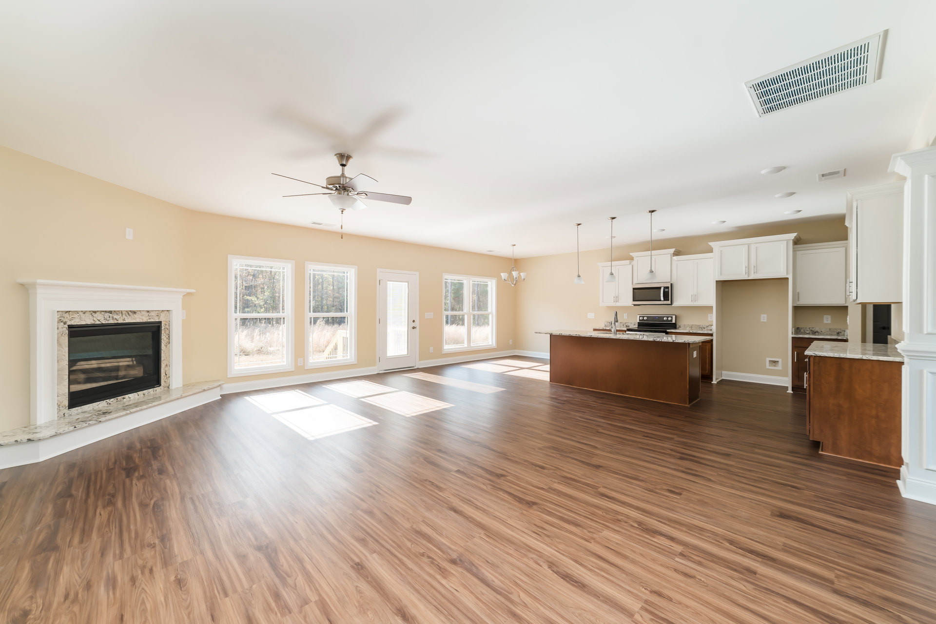 Spacious living room with hardwood floors, stone fireplace with glass doors, open kitchen featuring white cabinetry, sink, and faucet, large windows revealing trees outside