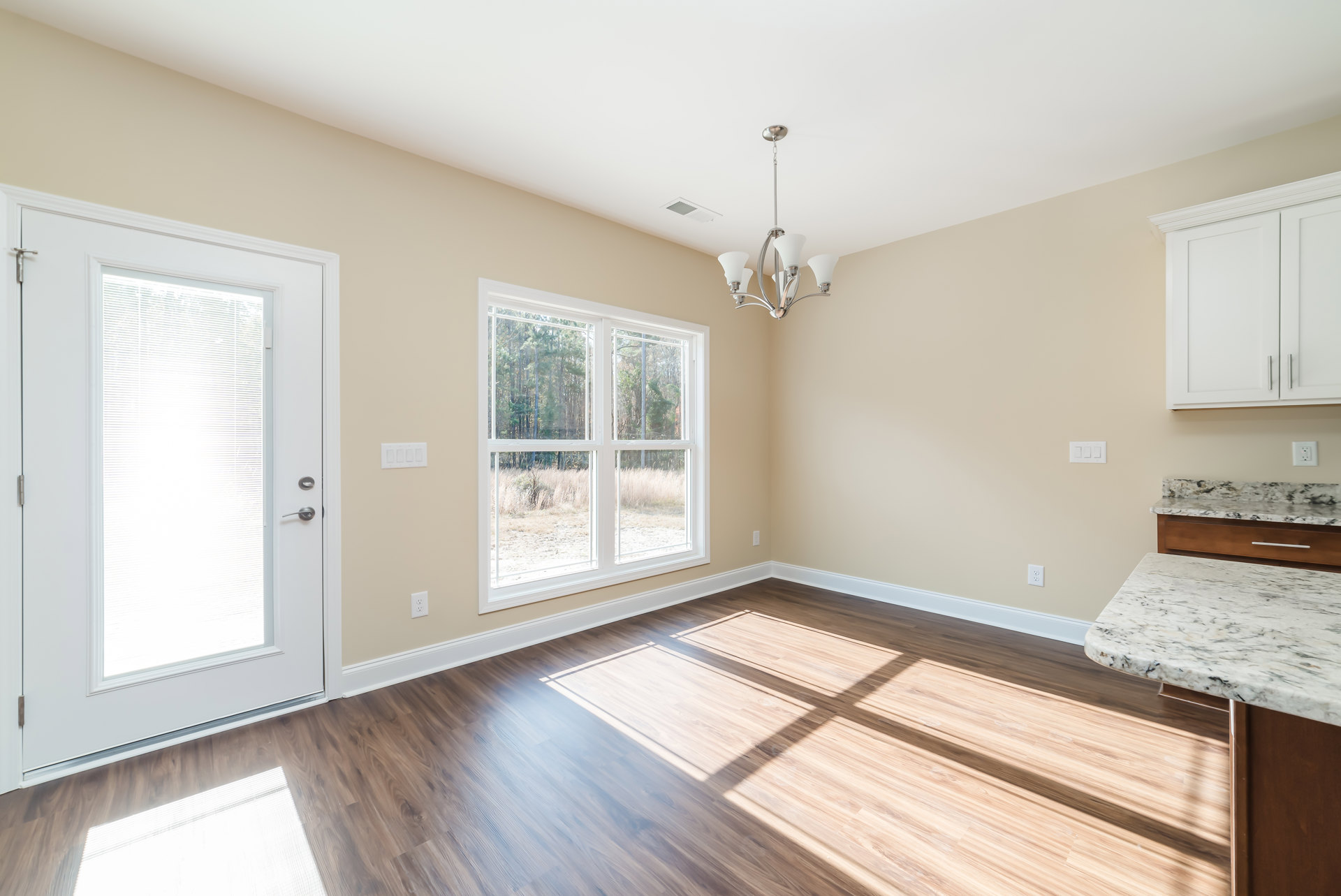 Open kitchen with wood flooring, white cabinetry with silver handles, marble countertop, large window overlooking trees, and white door with blinds.