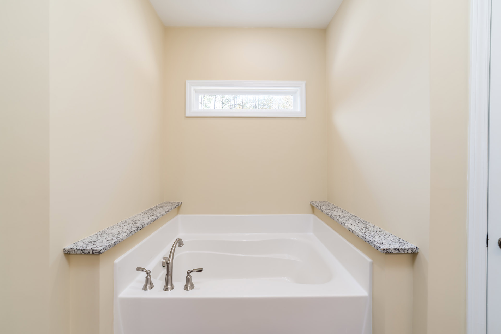 White freestanding bathtub with chrome faucet beside granite countertop, large window overlooking trees, light tile walls and floor, modern bathroom fixtures