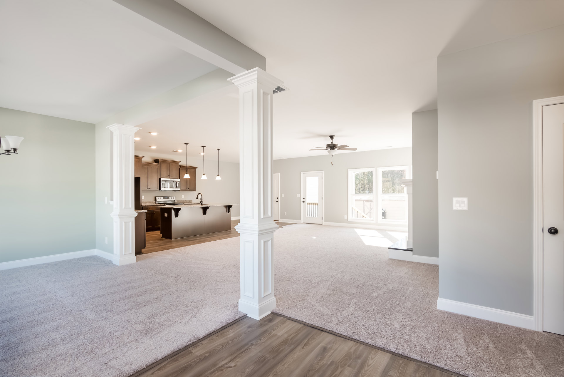Spacious room with white columns, carpeted floor, ceiling fan, and adjacent kitchen and dining area; white door with glass panels and metal railing visible.