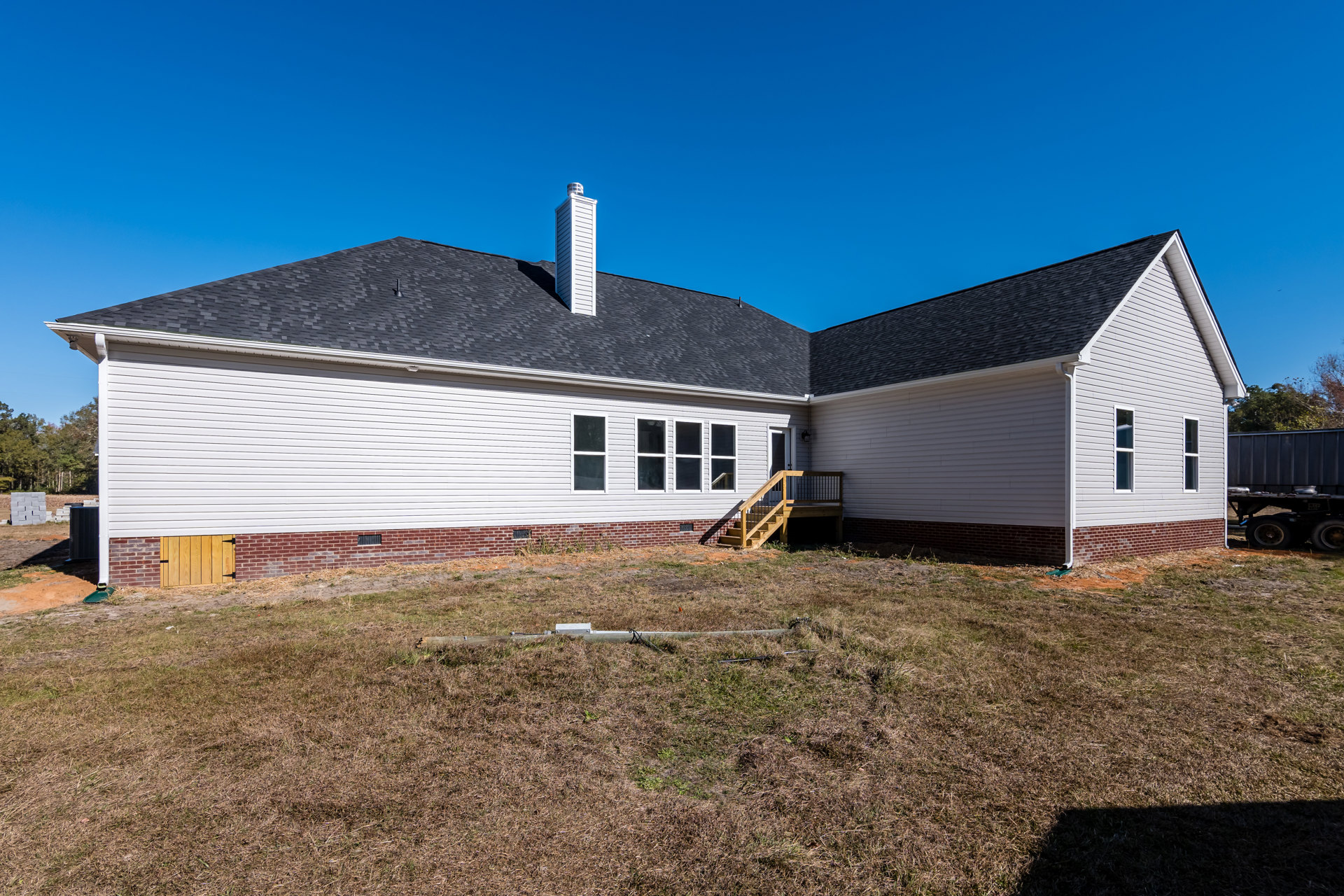 Two-story cottage with white siding, prominent white chimney, wooden exterior staircase, grassy yard, and metal trailer beside the house