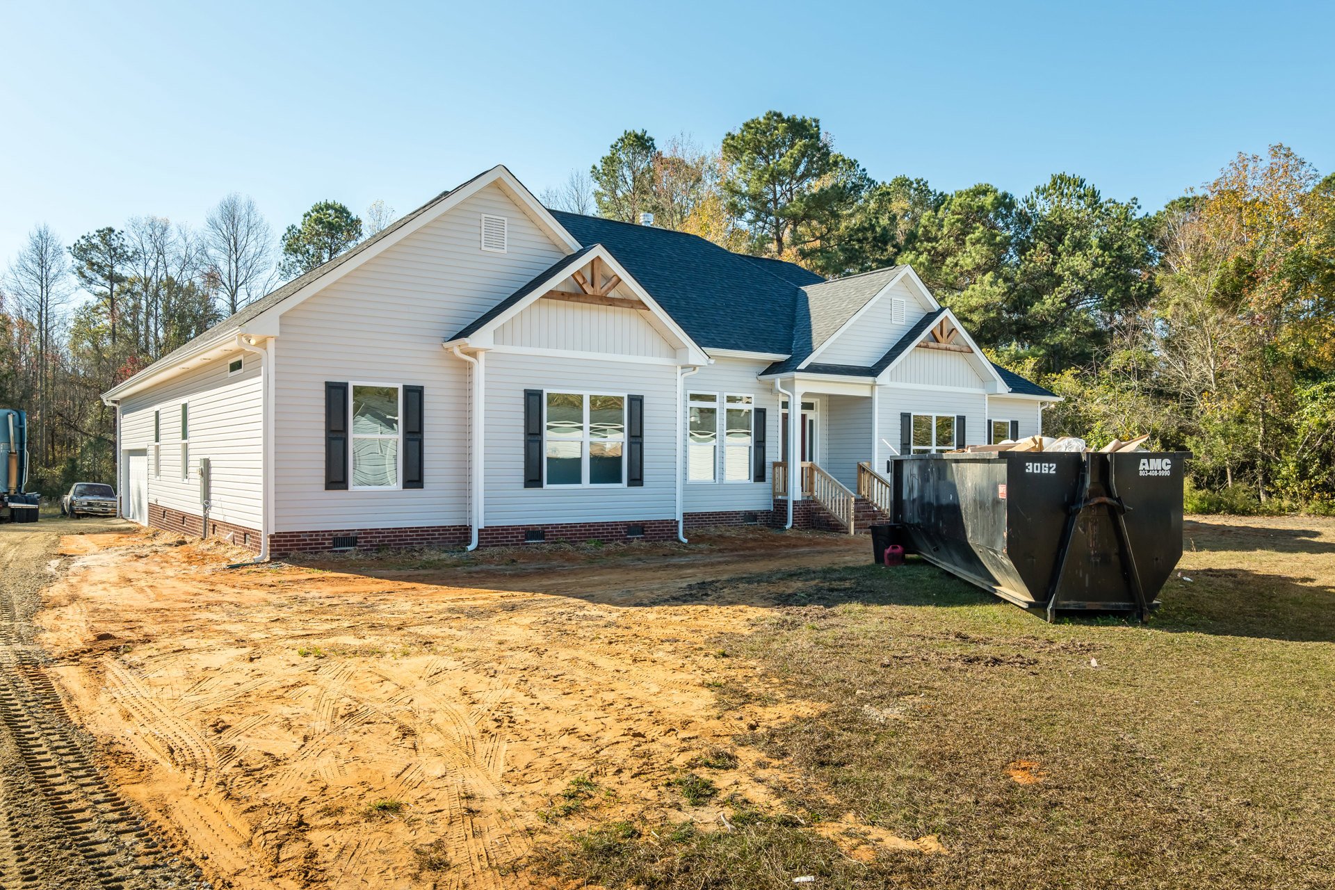 Black-roofed house with white-framed windows, black shutters, and a large black dumpster in front; white chair visible inside window, trees and sky in background