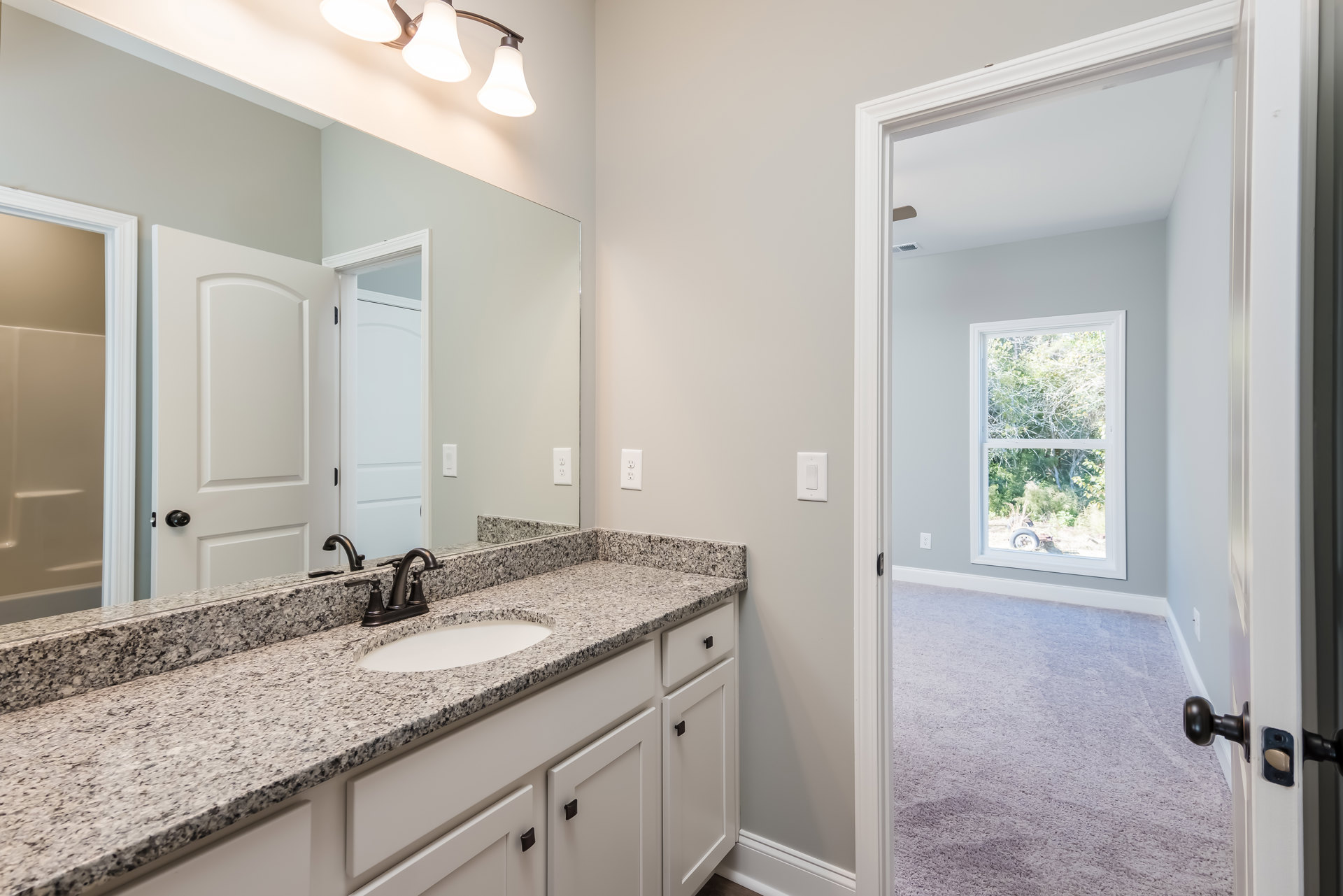 Marble bathroom countertop with undermount sink and chrome faucet, large framed mirror above, white cabinetry and door, window revealing leafy trees outside, modern light fixture