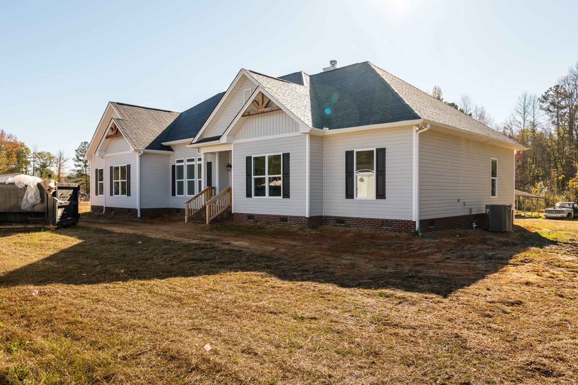 Two-story house under construction with exposed wooden porch, white-framed windows, black shutters, wooden staircase railing, unfinished yard, and blue sky overhead; white car