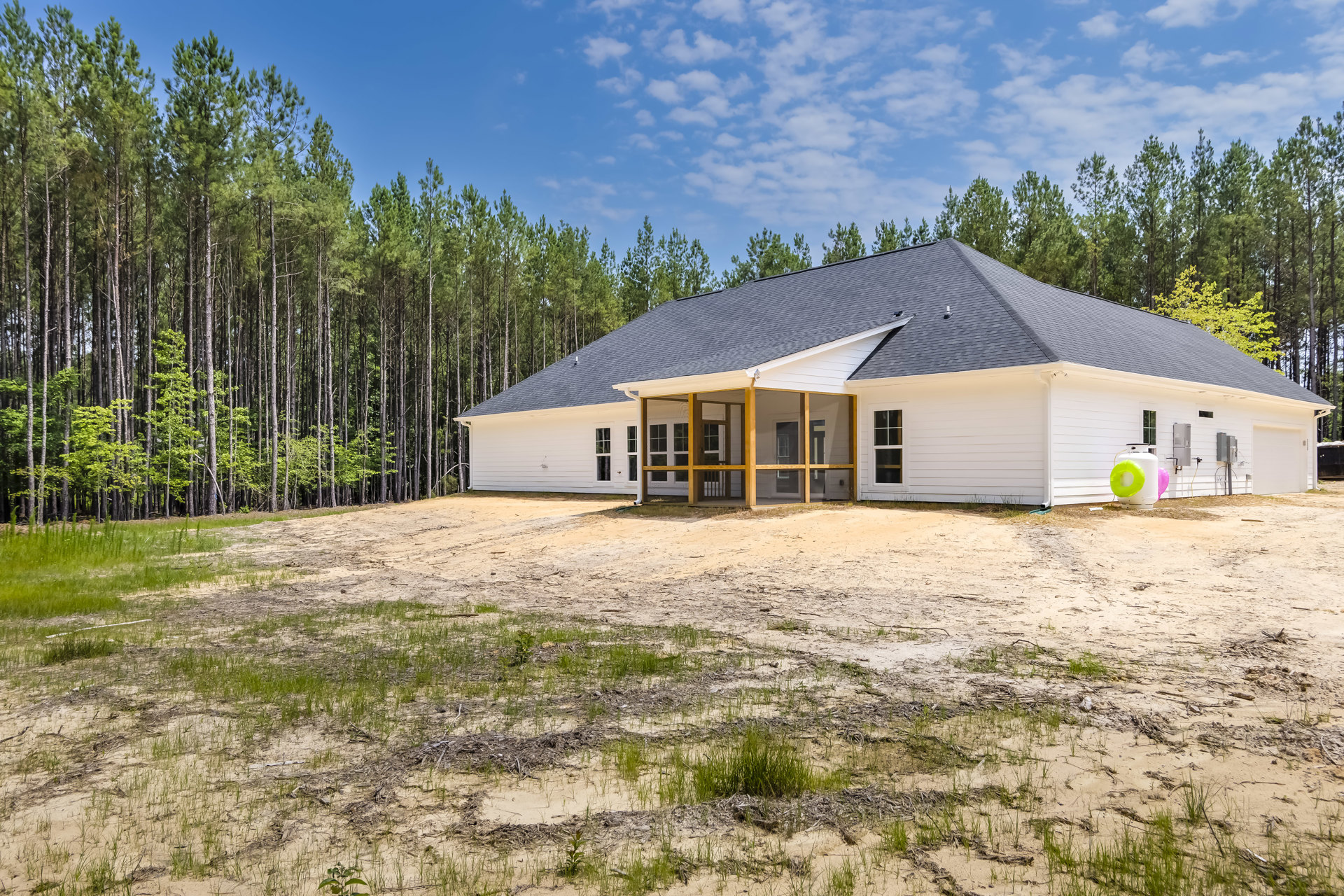 White siding house with black shingle roof, spacious grassy yard, large covered porch, screen door, mature trees in background under blue sky