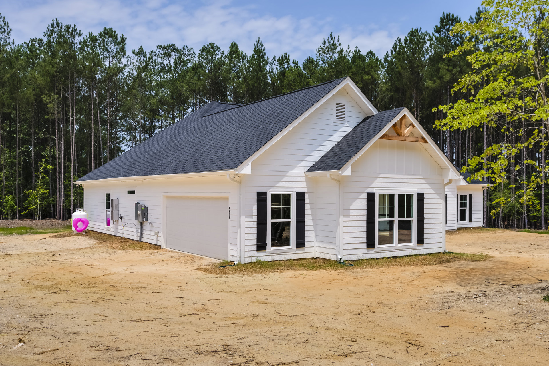 White siding house with black shutters and black roof, surrounded by green trees, under partly cloudy sky