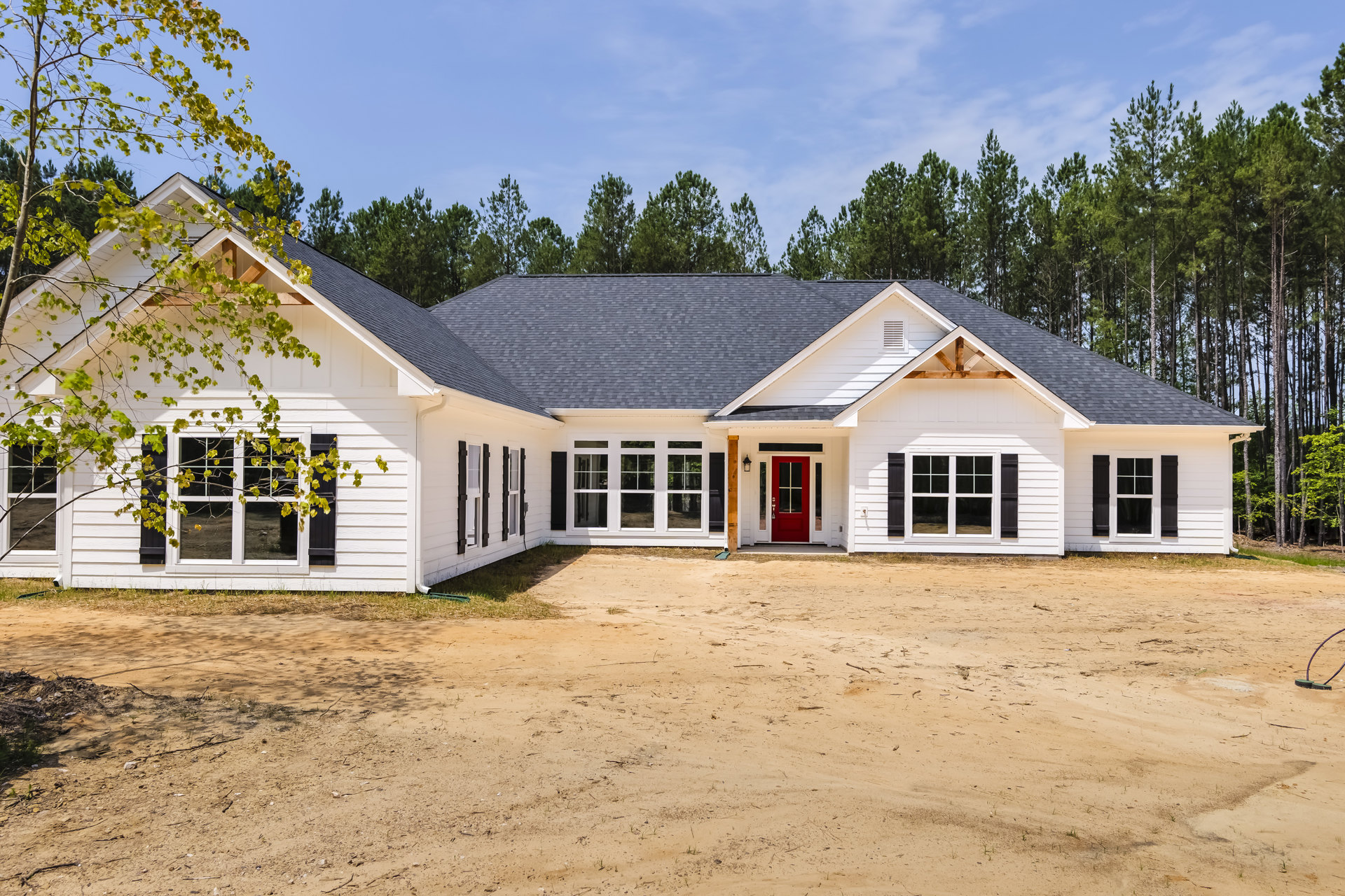 White siding house with a prominent red front door, black-trimmed windows, and a dirt yard under a partly cloudy sky.