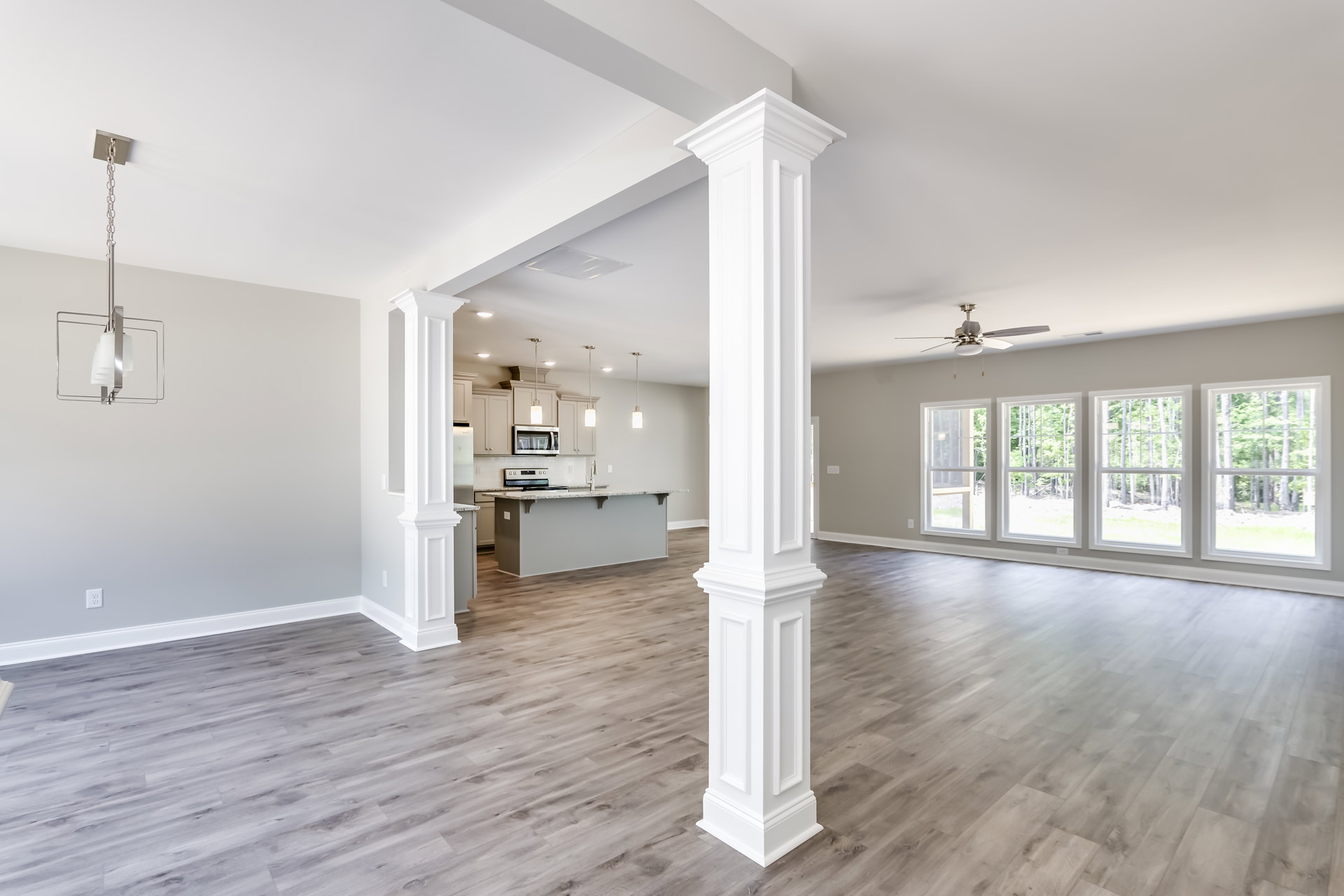 Open-plan kitchen and living area featuring a prominent white column, white paneled door, wood flooring, white-trimmed windows with views of trees, and a metal pendant light