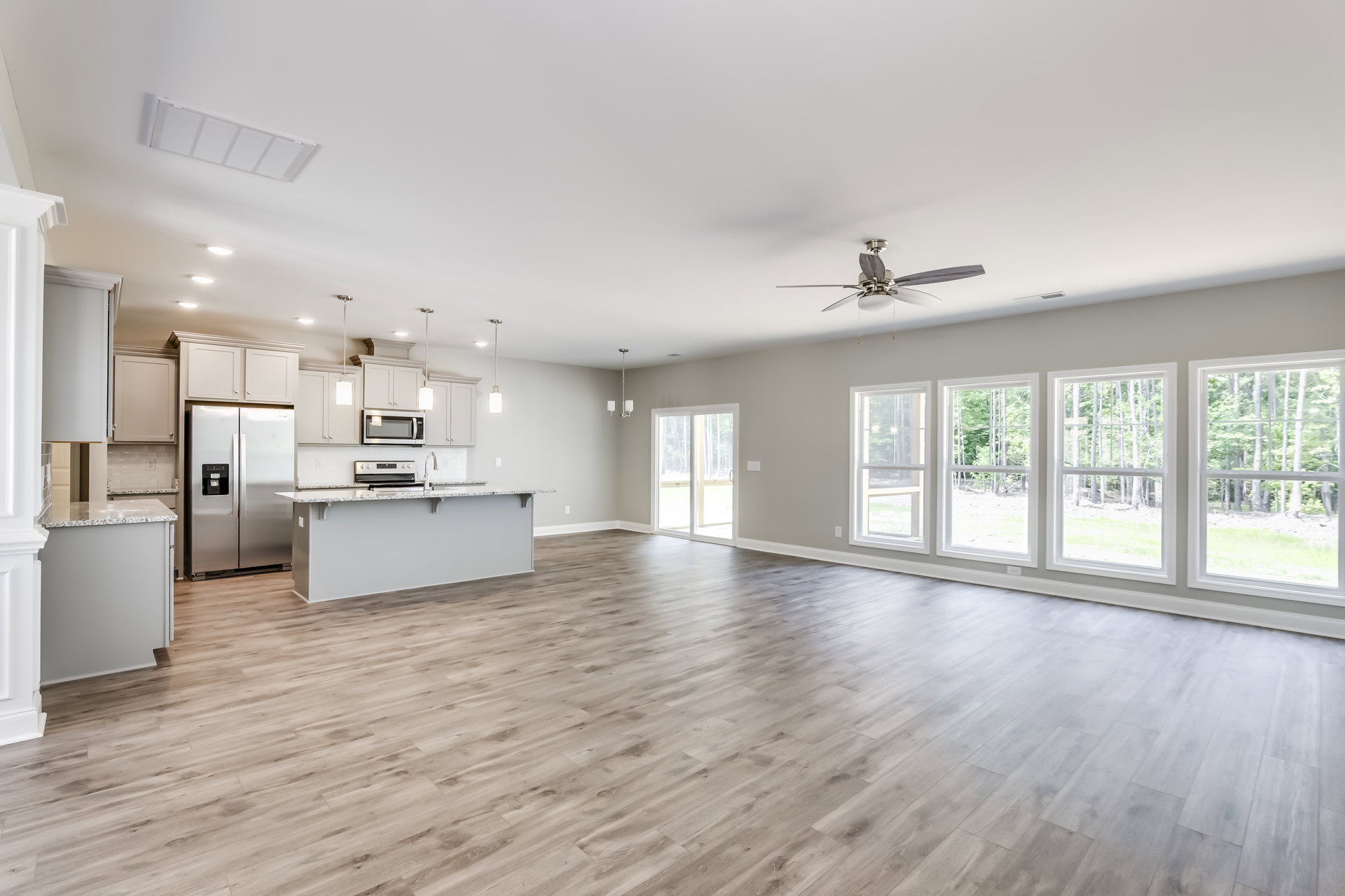 Spacious open floor plan featuring hardwood flooring, central kitchen island with light countertops, stainless steel refrigerator, built-in microwave, white cabinetry, ceiling fan