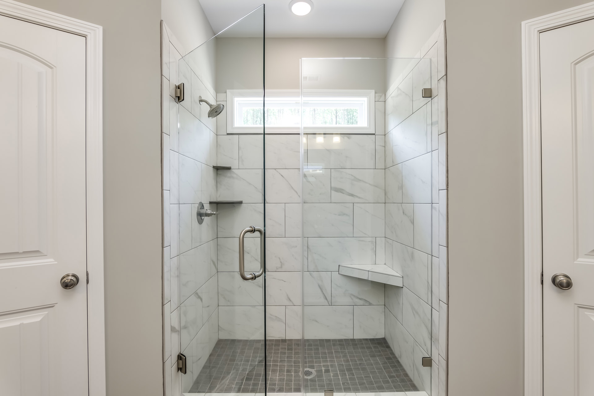 Glass shower enclosure with window, white tiled walls featuring a black accent line, metal door handle and towel bar, floor drain visible, natural light entering through window
