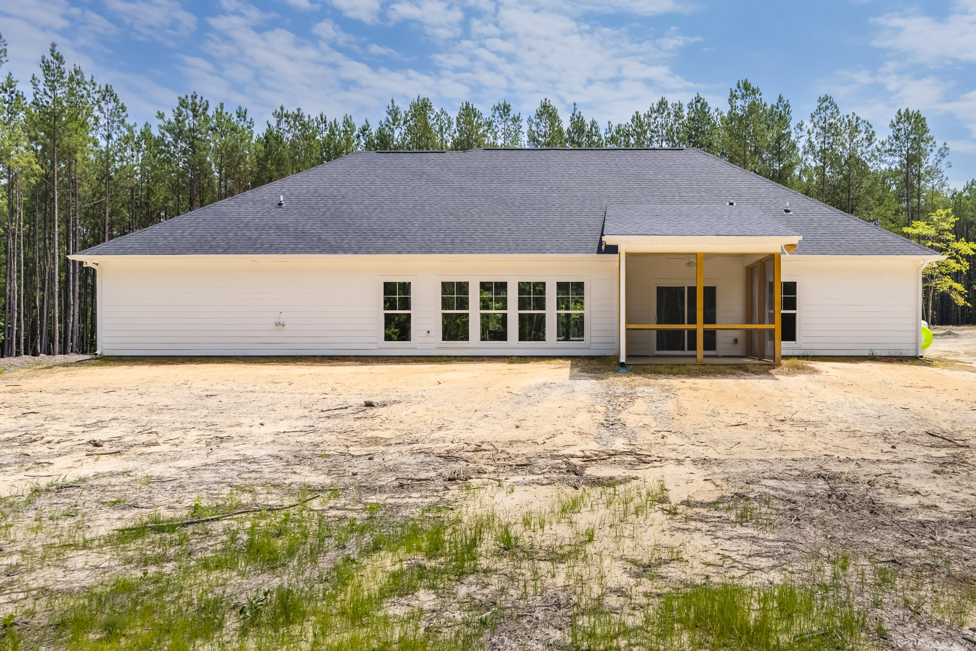 Two-story house with screened porch, multiple white-framed windows, and unfinished dirt yard surrounded by trees under a cloudy sky