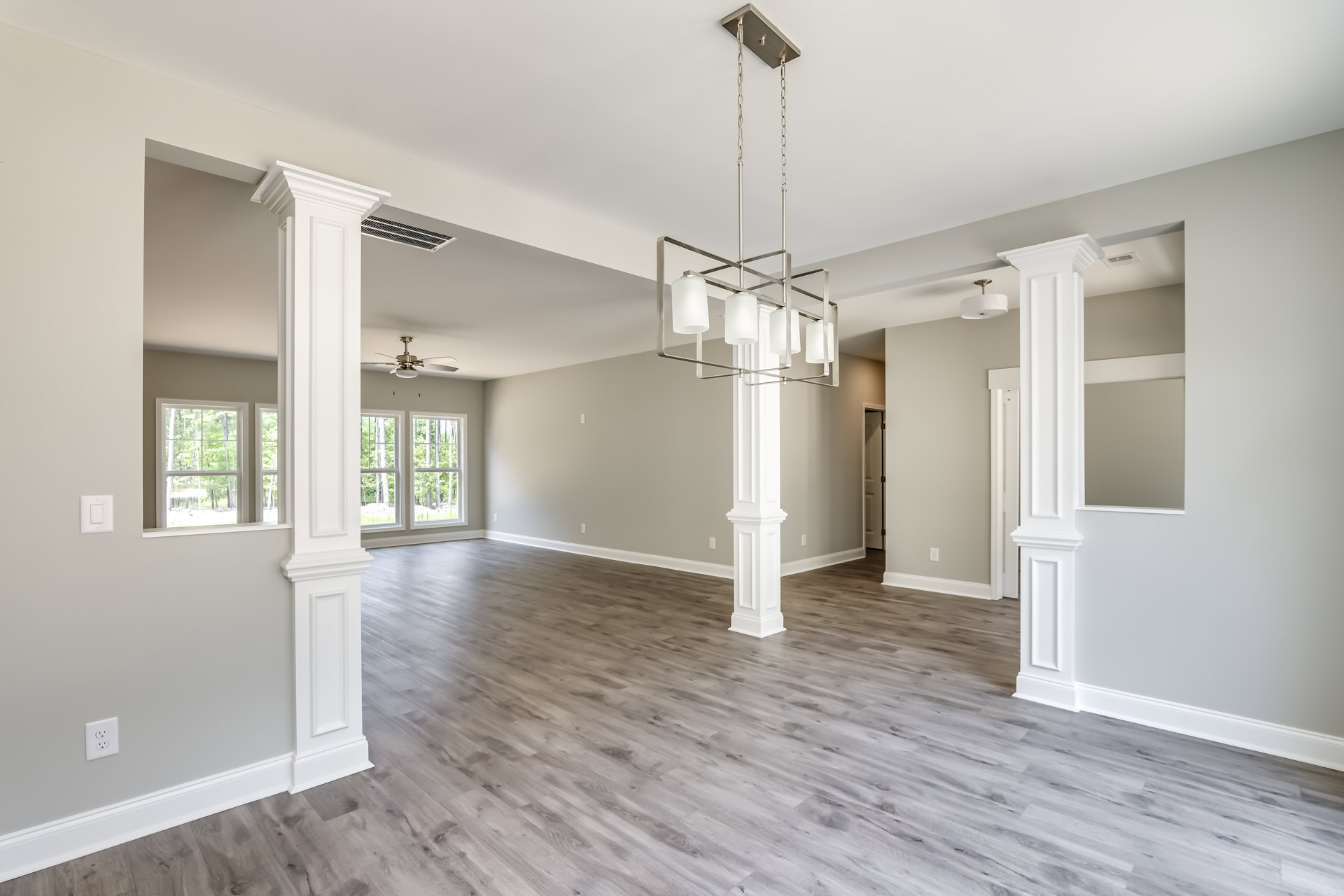 Spacious living area with white columns, ornate chandelier, wood laminate flooring, and large window overlooking trees