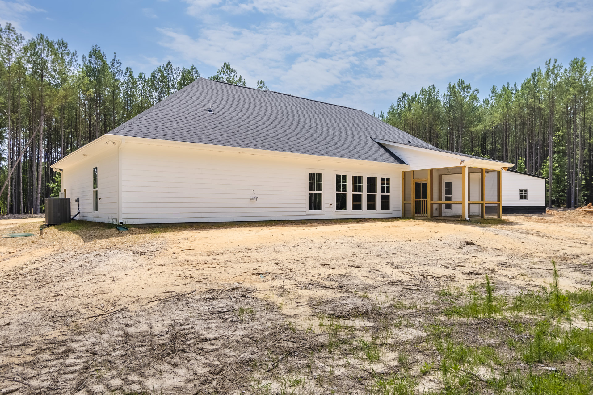 White siding house with covered front porch, black framed windows, metal HVAC unit on side, surrounded by grass, dirt driveway, and trees under blue sky