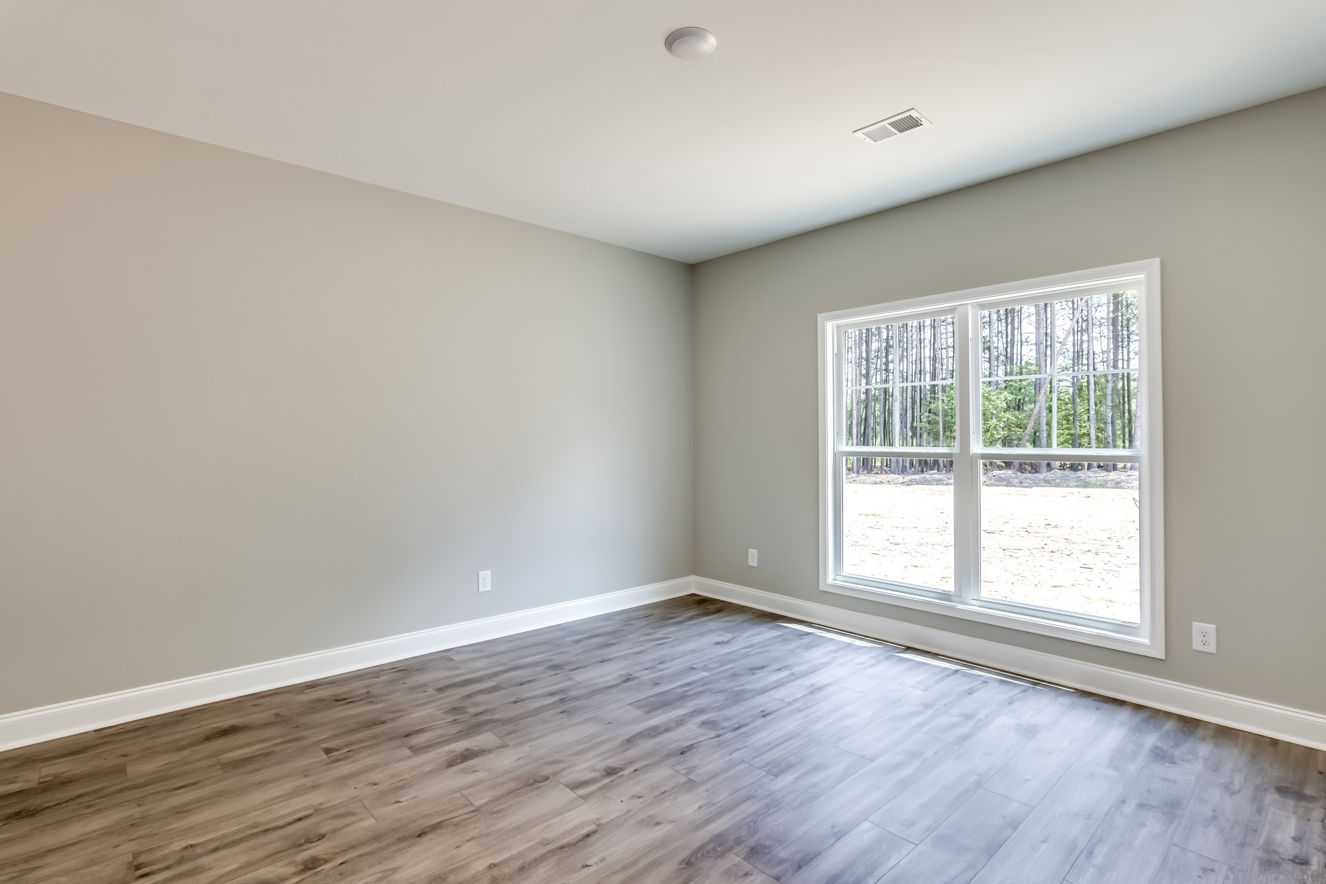 Sunlit room with large window overlooking trees, wood laminate flooring, white plaster walls, ceiling vent, and round recessed light fixture