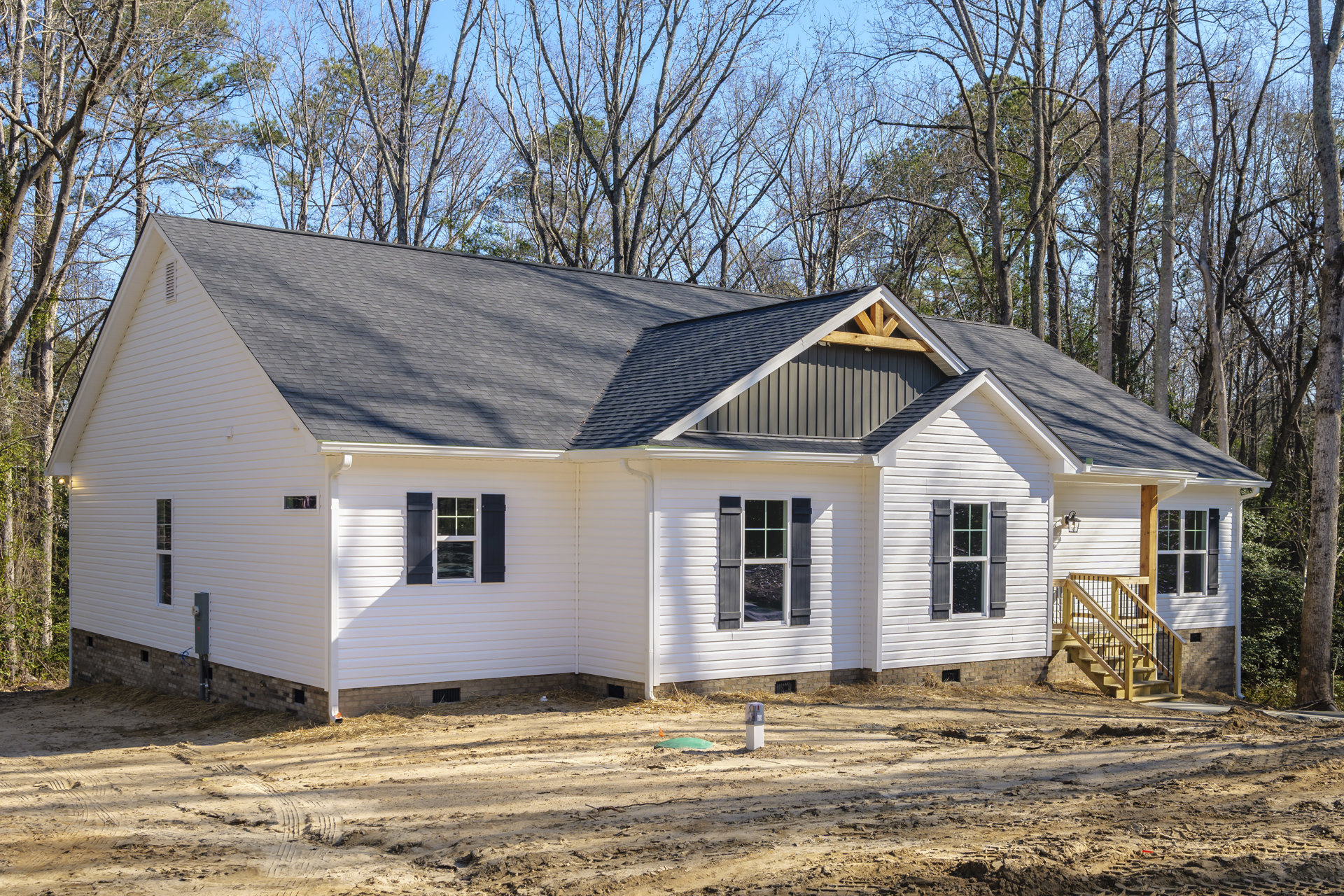 Two-story house under construction with exposed wooden stairs, white-framed windows, black shutters, and a dirt path leading to the entrance