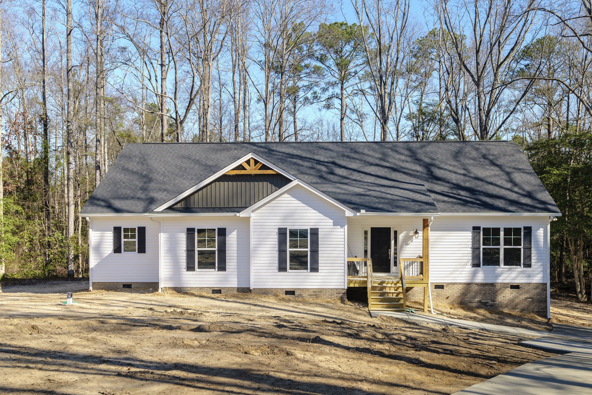Two-story house with light siding, front porch, wooden stairs, cross-mullioned window, dirt path leading to entrance, surrounded by trees under a clear sky
