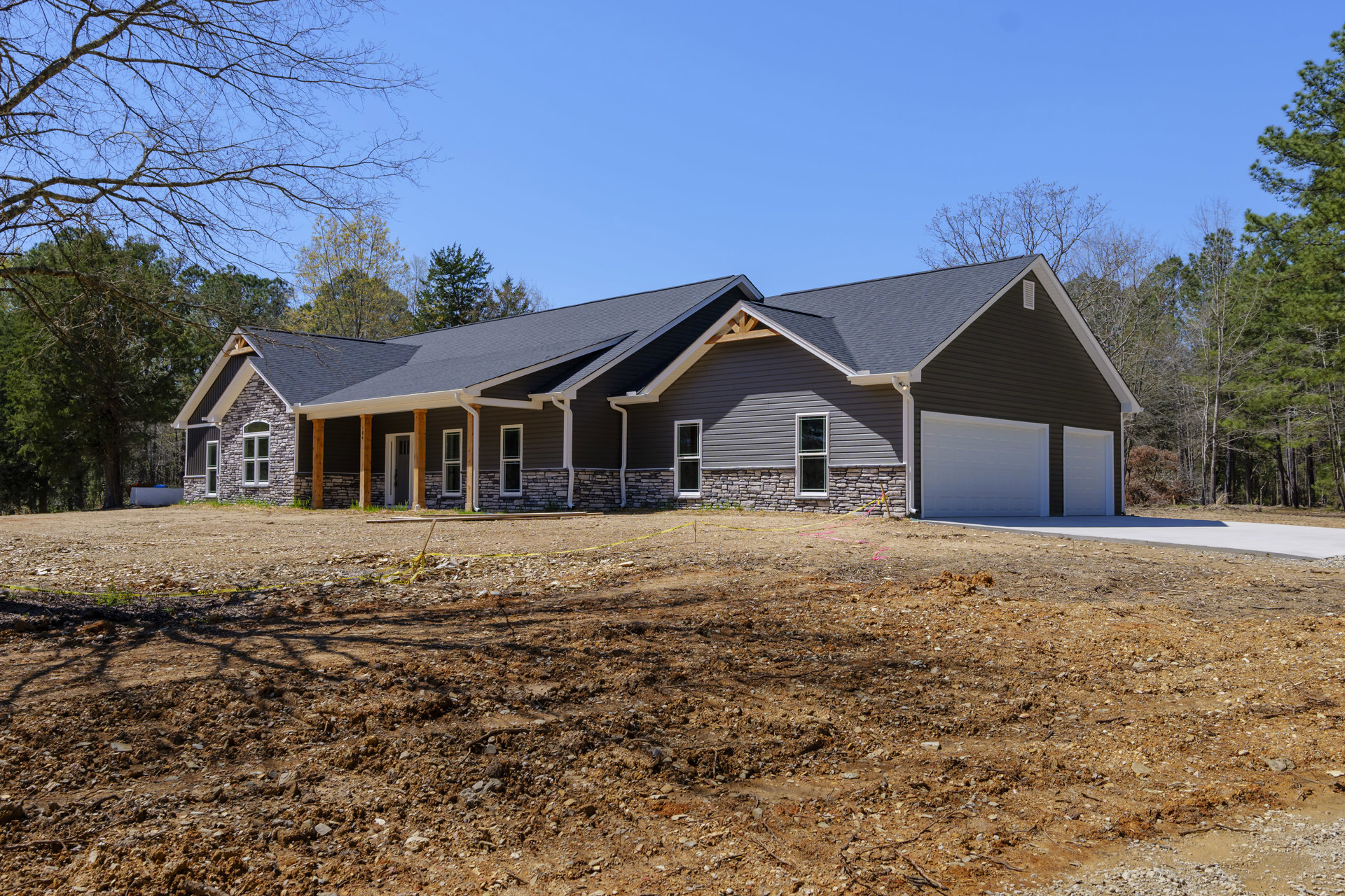 White siding house with black trim, stone accent wall, attached garage with white door, paved driveway, trees behind roof, dirt ground in foreground