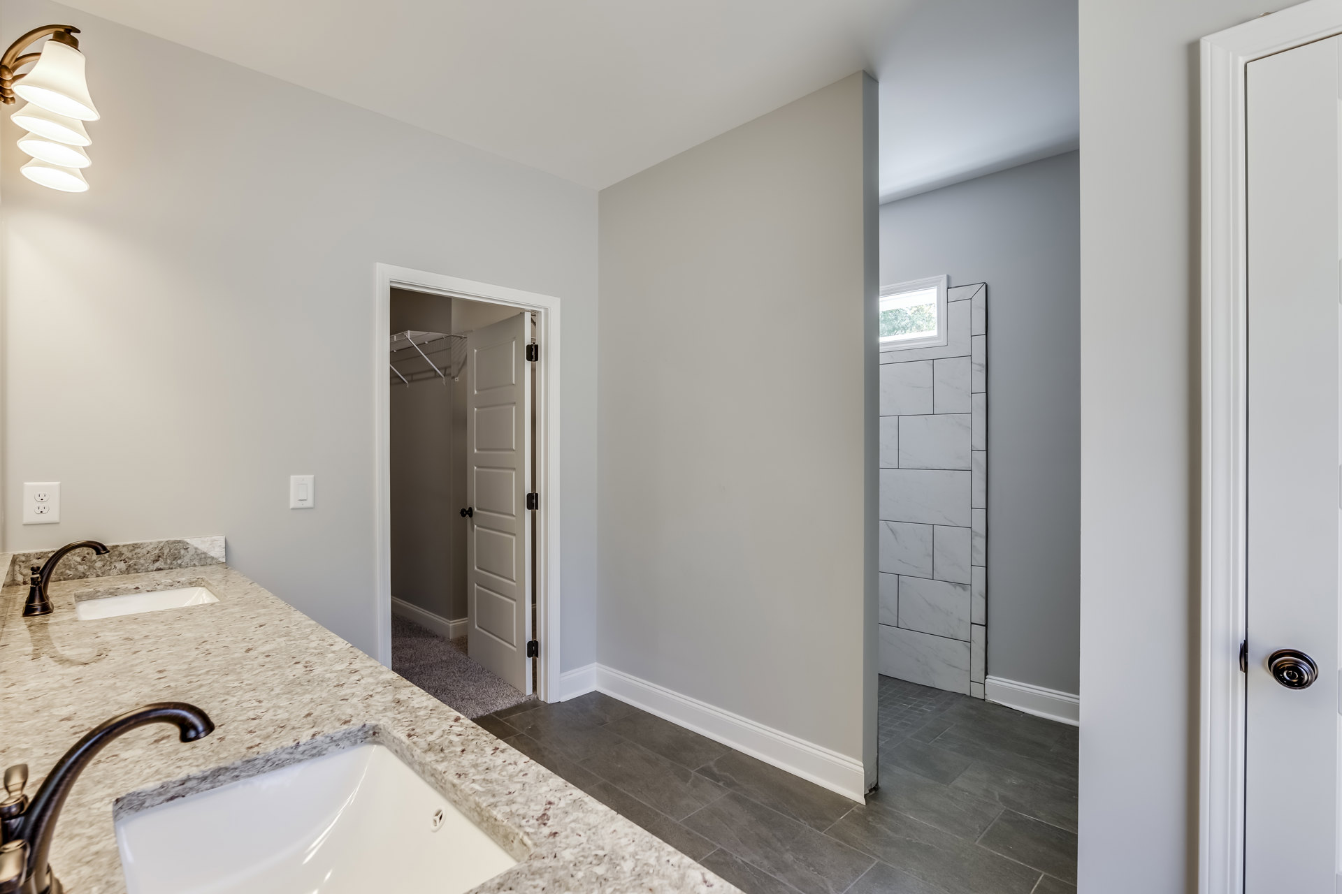 White bathroom featuring a marble countertop sink, chrome faucet, wall-mounted lamp, frameless mirror, glass shower enclosure, and white tile walls with black trim door.
