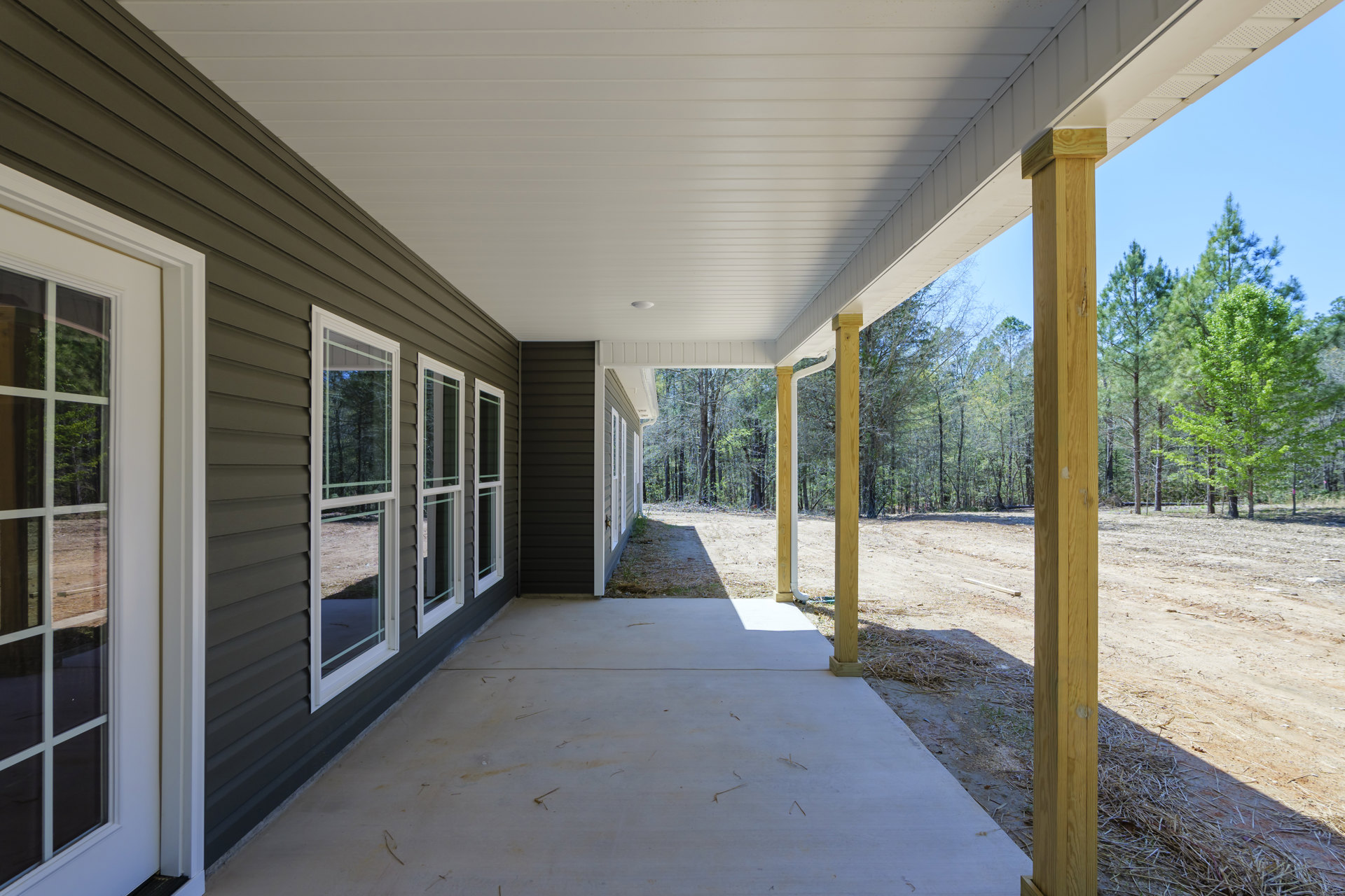 Covered porch with white ceiling and recessed light, row of windows overlooking wooded landscape and dirt road, concrete patio, exterior house door, surrounding trees