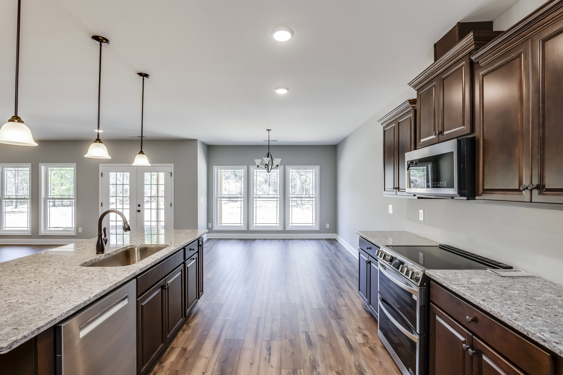 Kitchen with natural wood flooring, white cabinetry, stainless steel microwave, pendant chandelier, large windows overlooking trees, black accent pole on white countertop