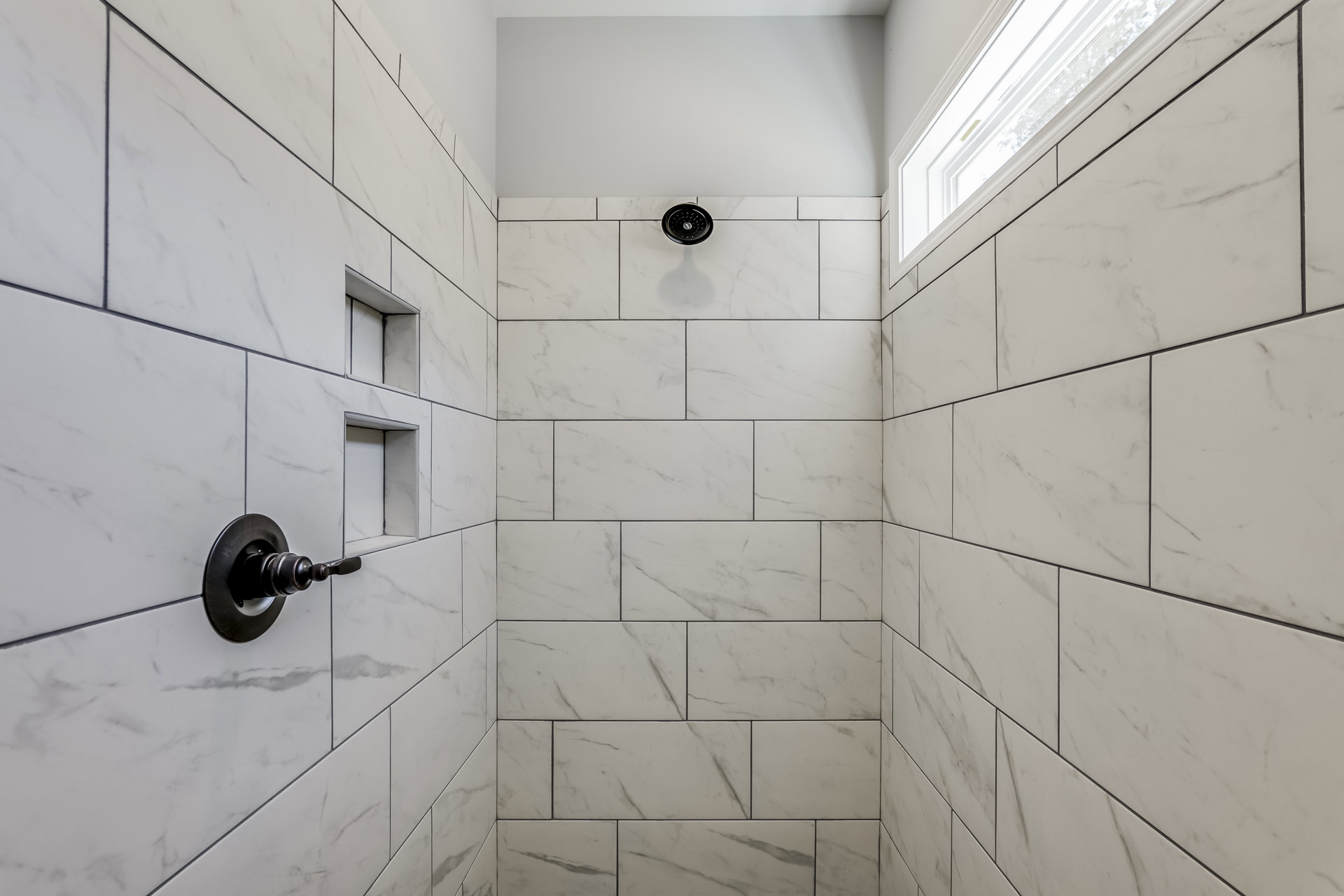 White tile shower wall with black round shower head, matching handle and knob, rectangular niche with black lines, and window letting in natural light