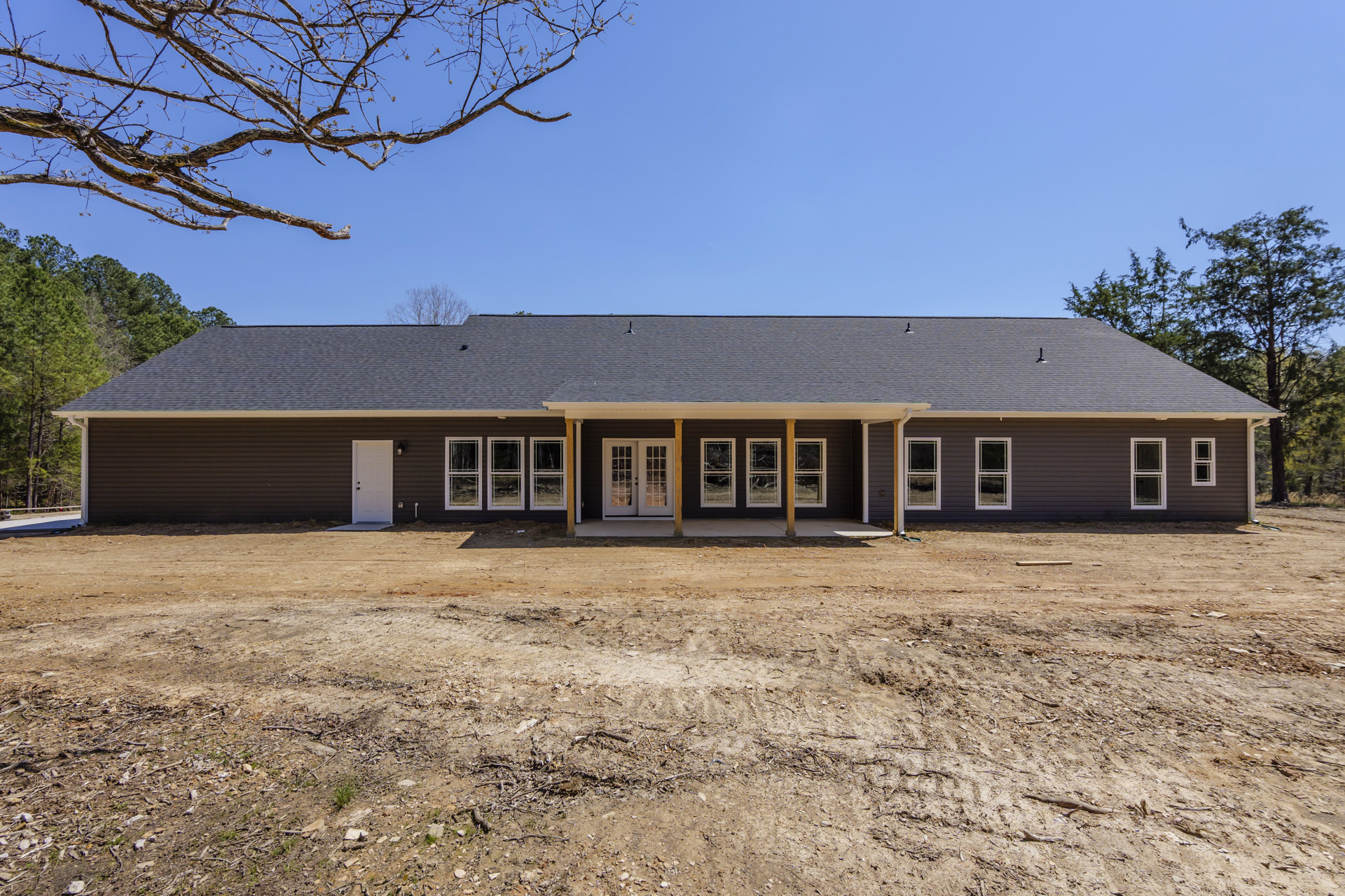 Two-story cottage-style home with white siding, double glass-paneled doors, multiple windows with white frames, covered front porch, expansive grassy yard, mature trees, and dirt