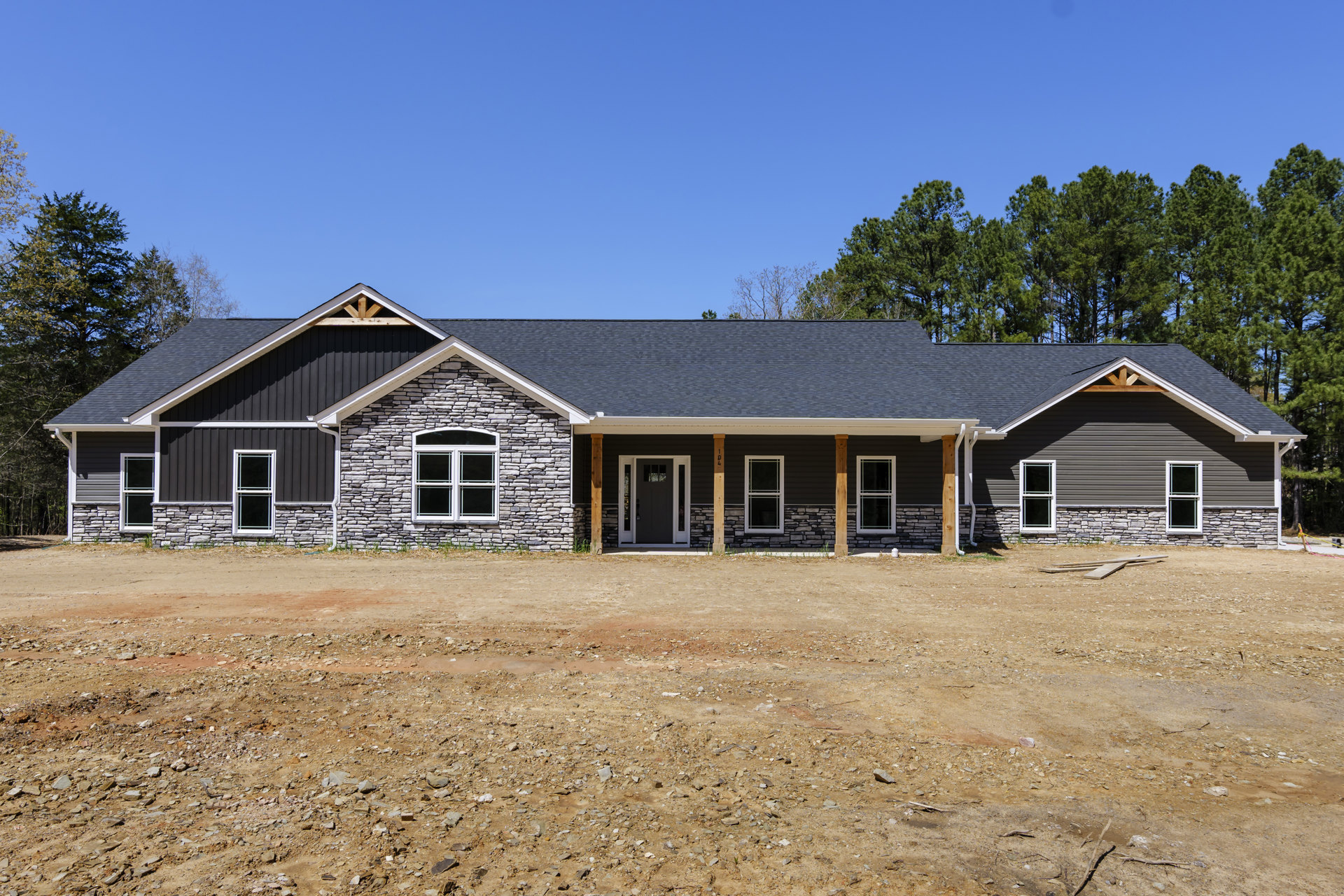 Partially built stone house with white-framed windows, black door, and stone pillars, surrounded by dirt ground and trees in the background