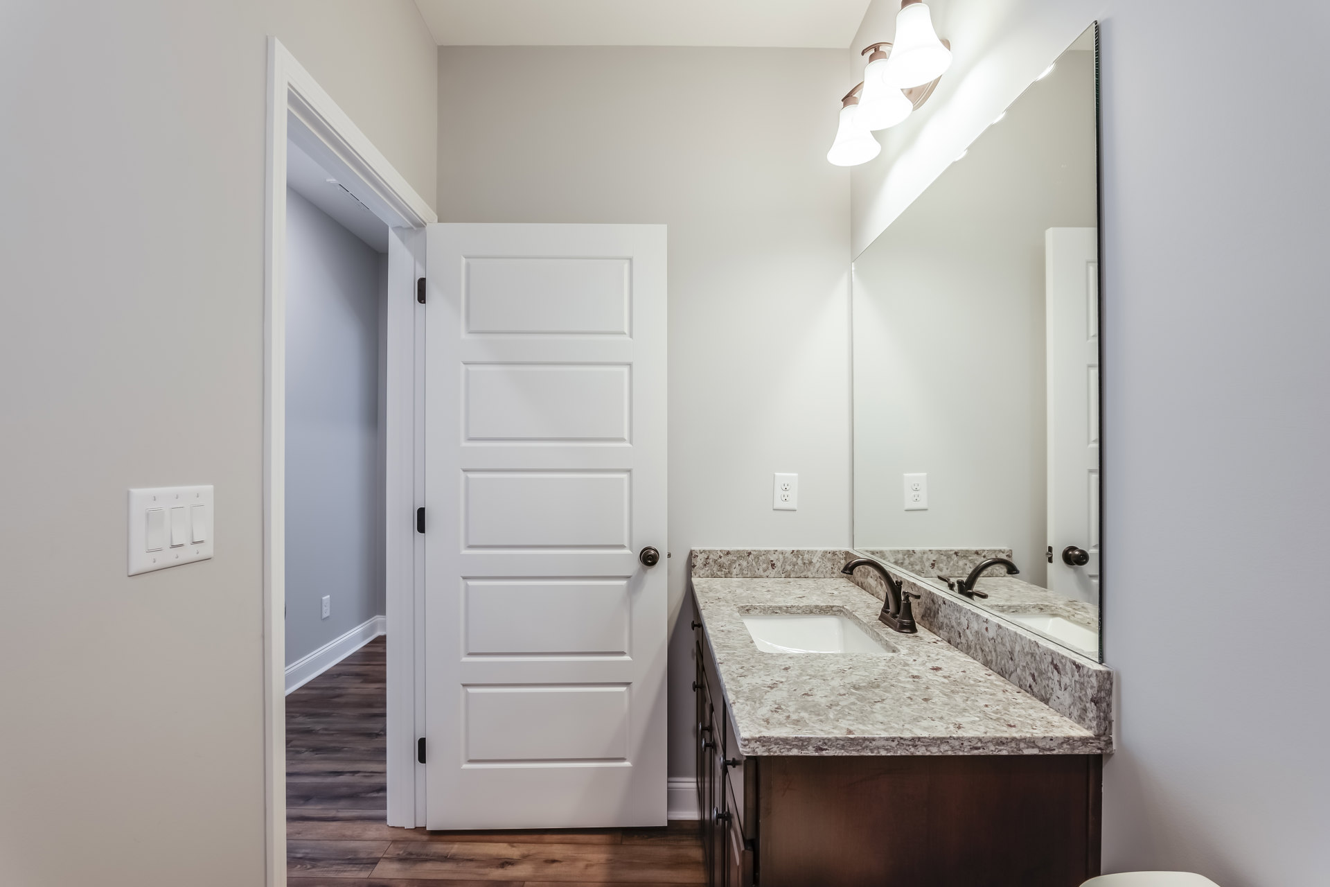 Bathroom with double sinks set in a marble countertop, wide mirror above, white cabinetry below, three-light fixture mounted on wall, white door with silver doorknob, close-up of