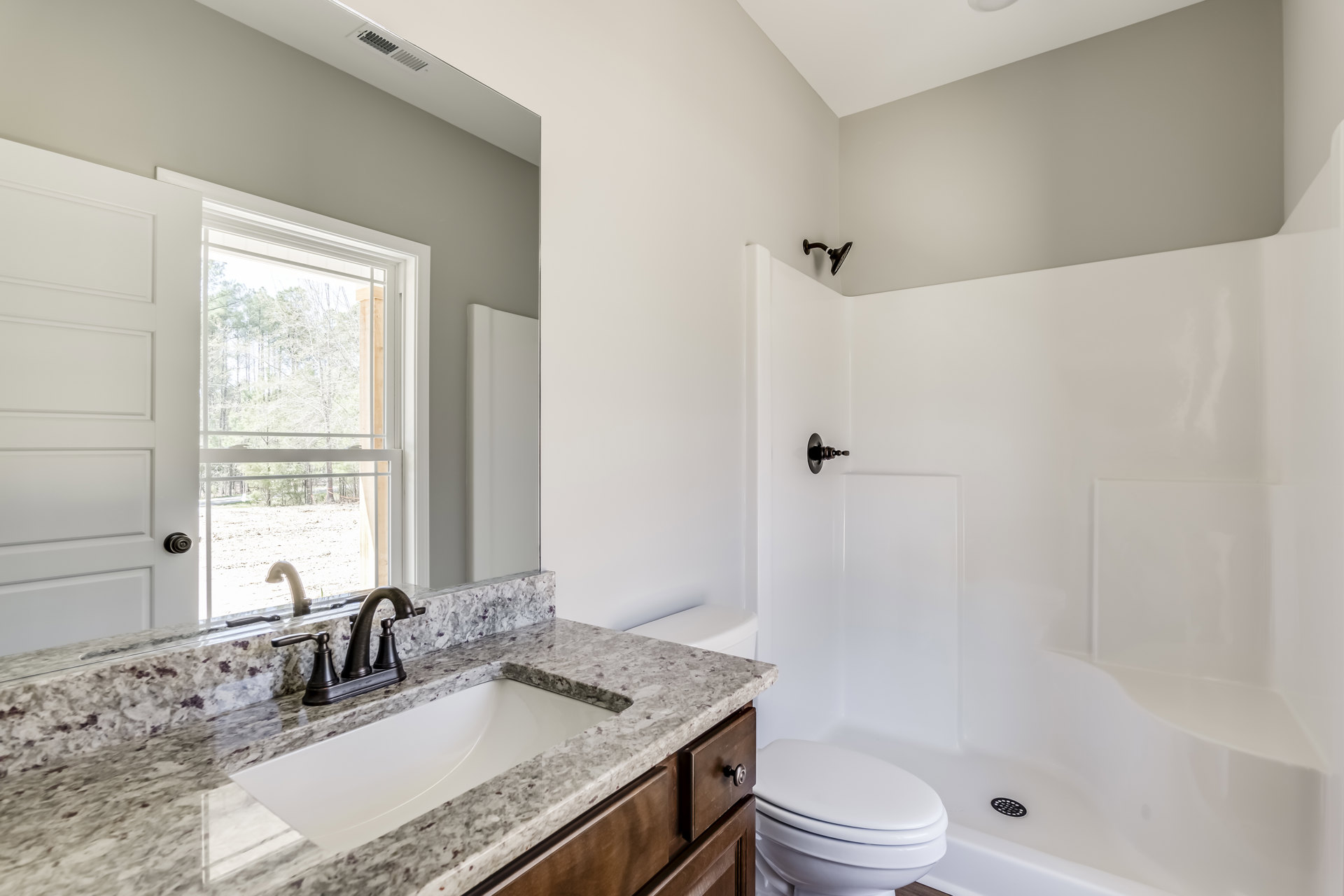 Marble countertop with black faucet, rectangular sink, white toilet, glass shower enclosure, large window overlooking trees, light tile flooring, white cabinetry