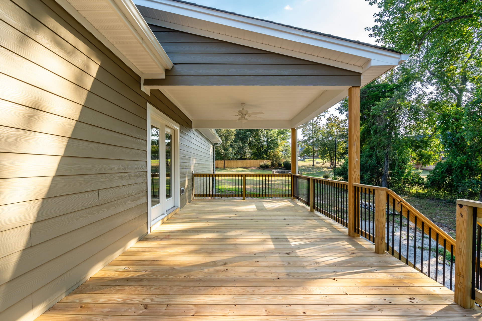 Wooden deck with black metal railing, covered porch featuring a ceiling fan, fenced yard, and leafy trees in the background.