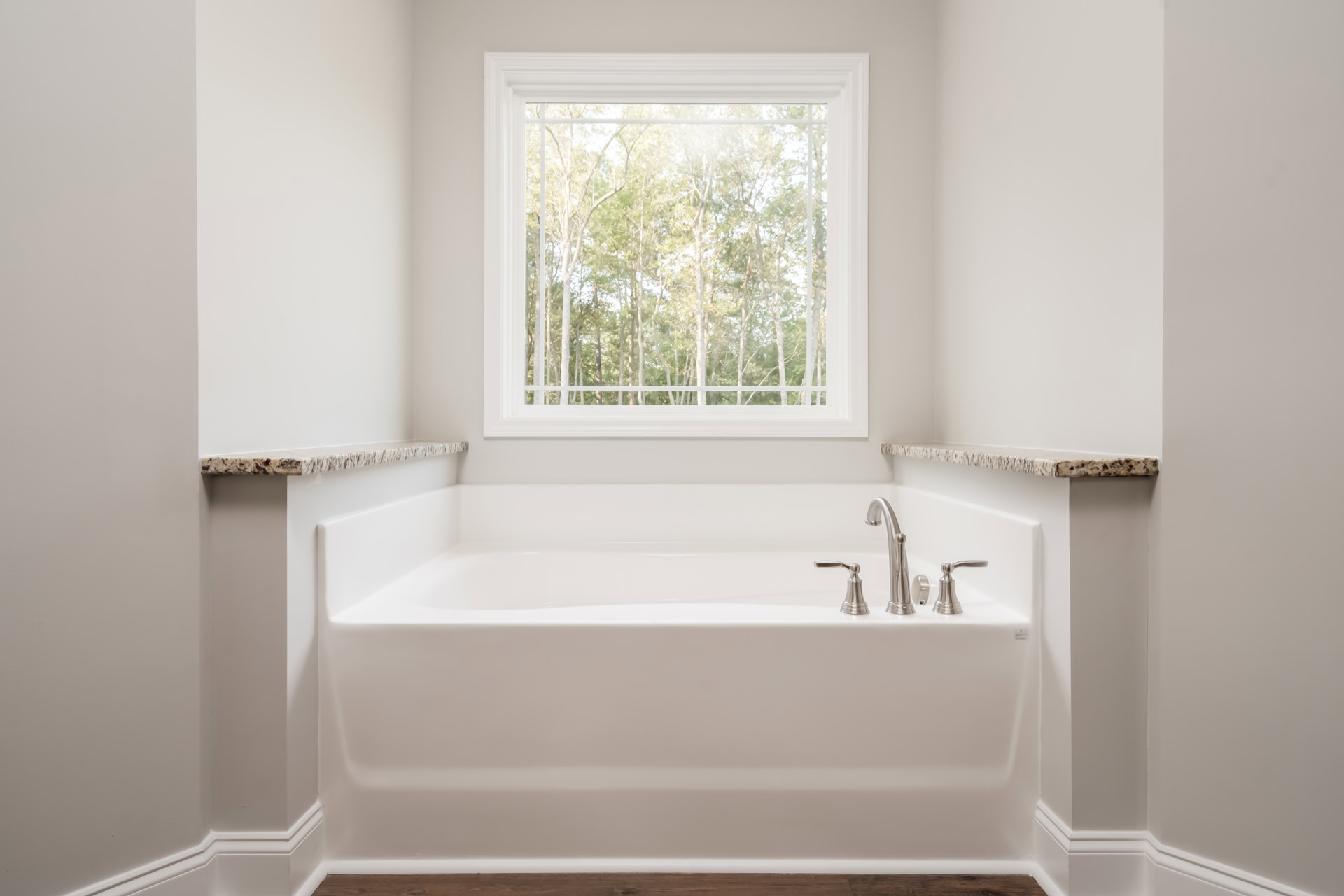 White freestanding bathtub beneath large window, chrome faucet, leafy trees visible outside, light-filled bathroom with neutral finishes