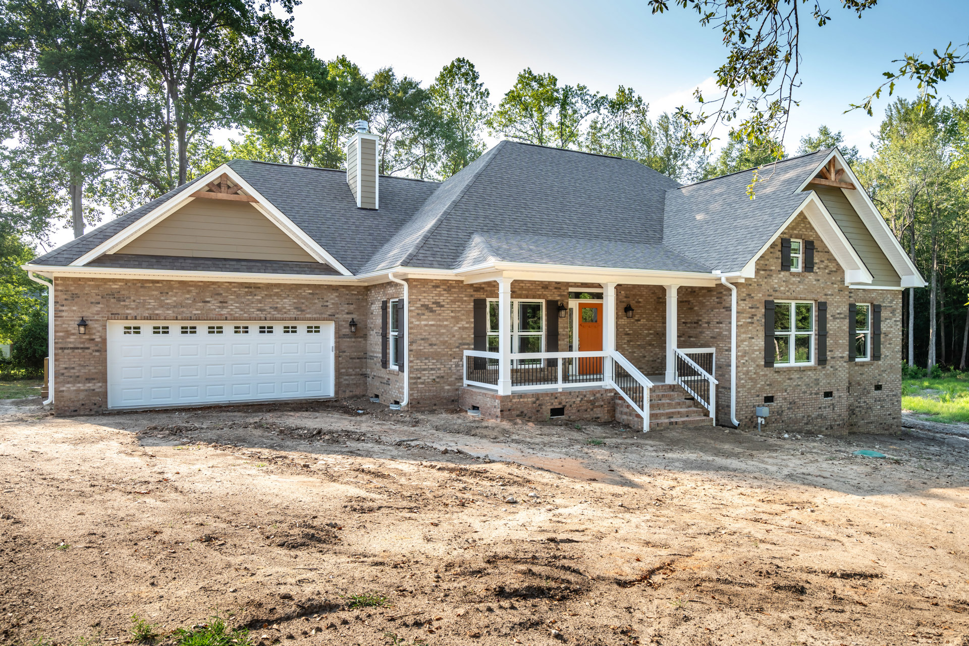 Two-story house with white garage door featuring windows, covered front porch with white entry door, brick chimney, and unfinished dirt driveway.
