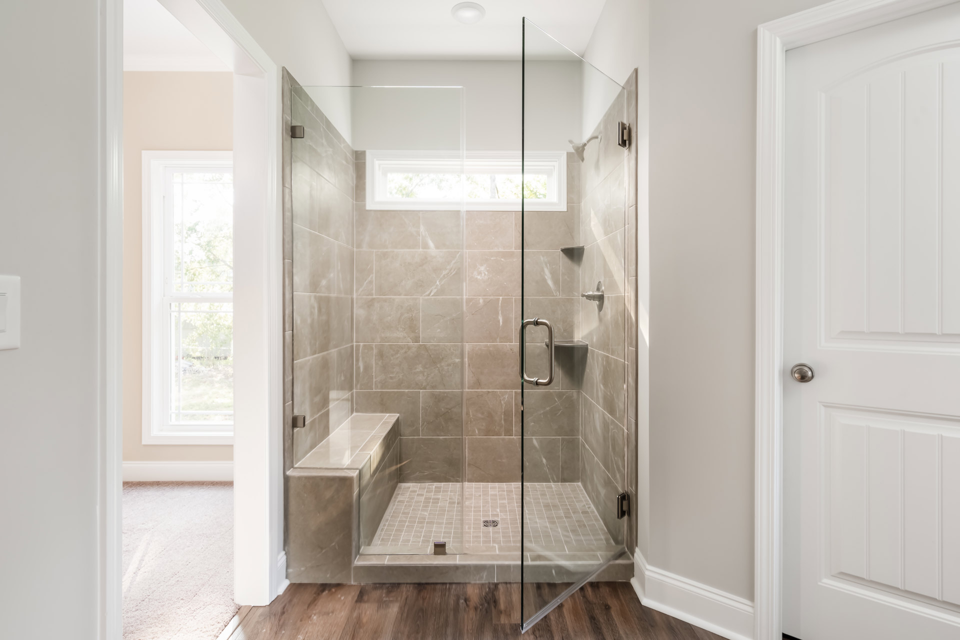 Glass shower enclosure with tiled walls, built-in bench, metal grab bar, white door with silver knob, window overlooking trees, light-colored flooring