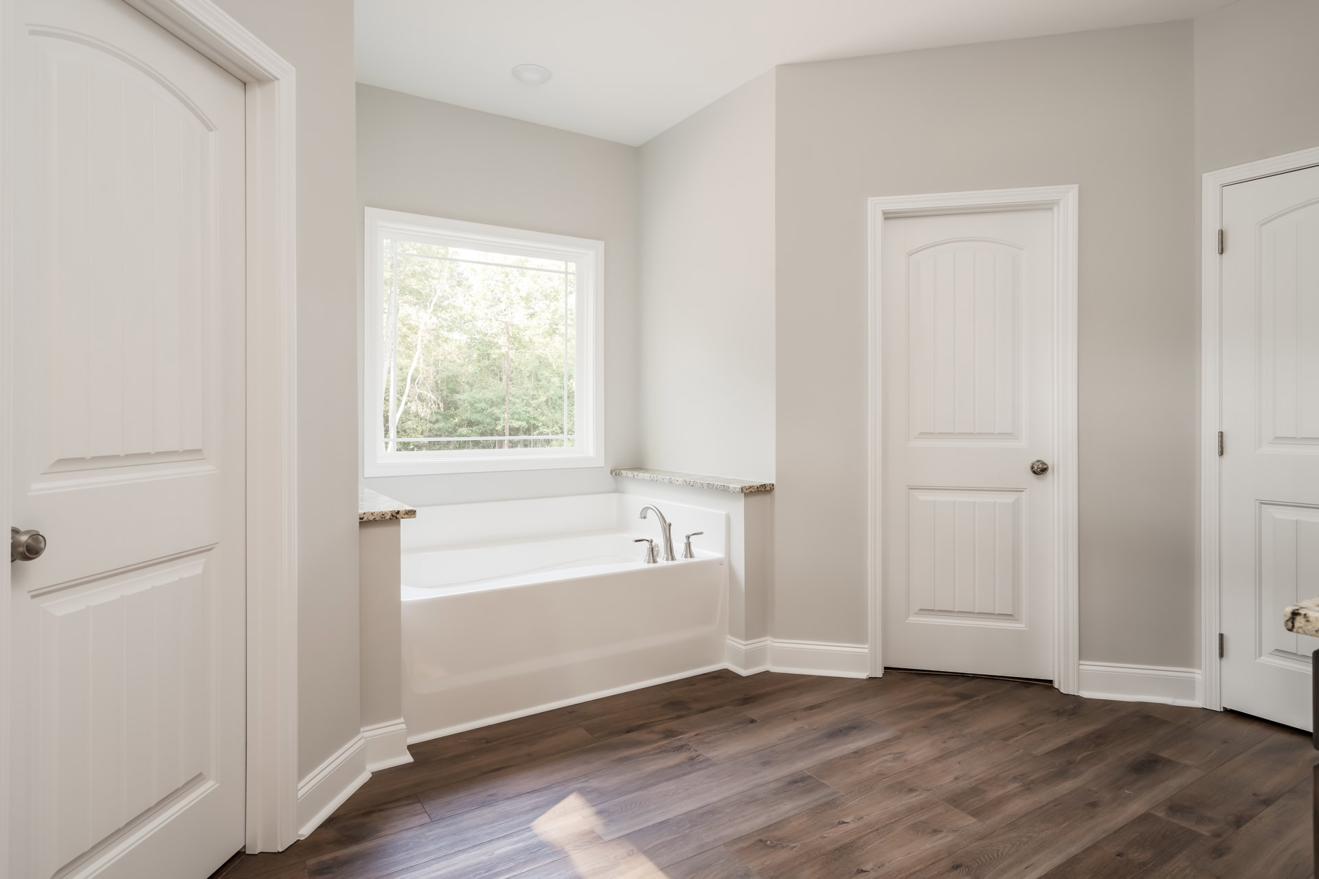 Bathroom with freestanding bathtub beside marble countertop, white walls, wood flooring, white door with silver knob, window overlooking trees