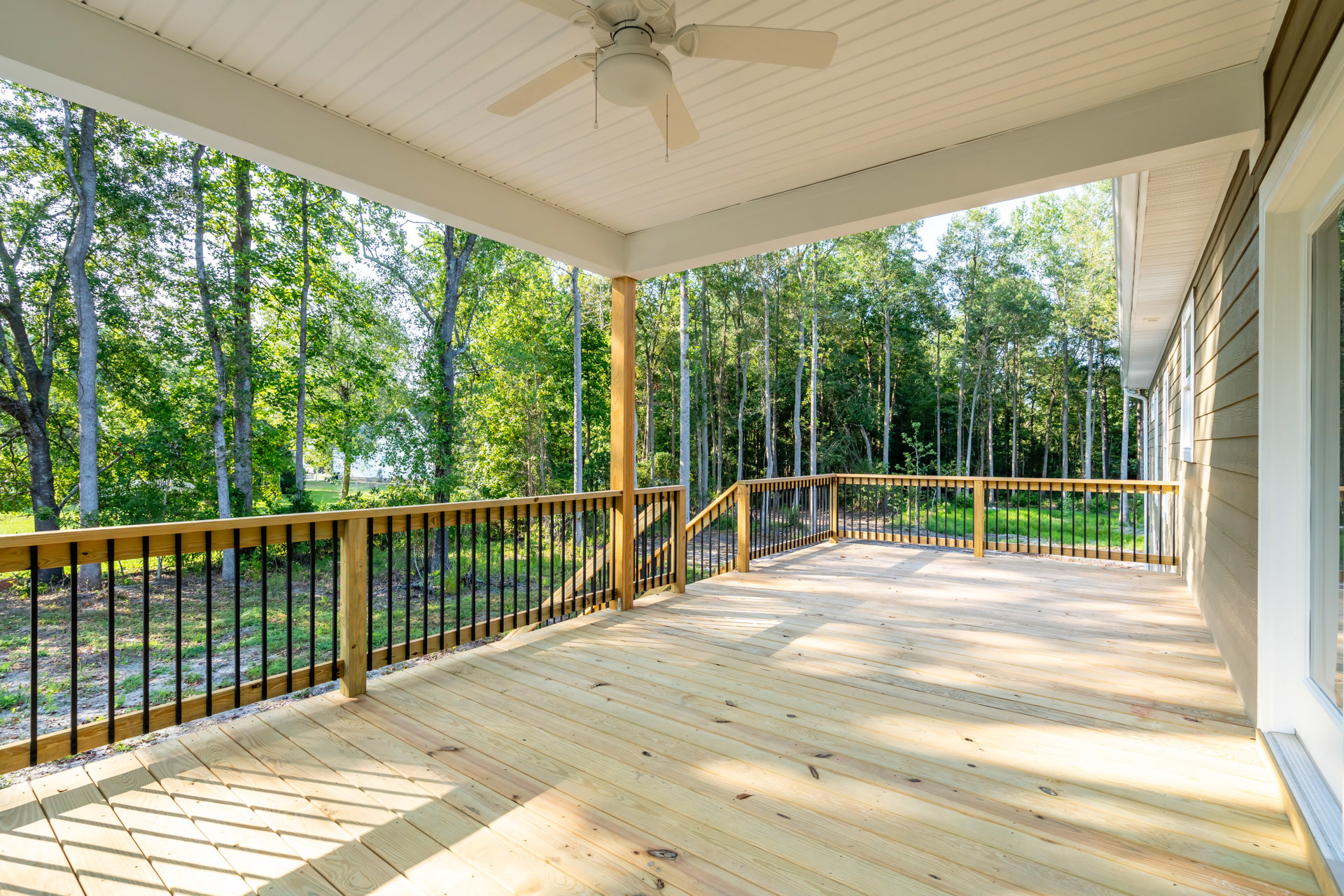 Wooden deck with black metal railings, ceiling fan overhead, surrounded by leafy trees in the background