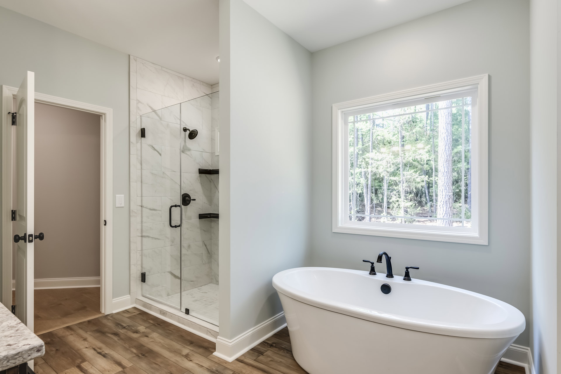 White freestanding bathtub with matte black faucets beside a glass-enclosed shower, light gray tile walls, large window overlooking trees, quartz countertop with undermount sink