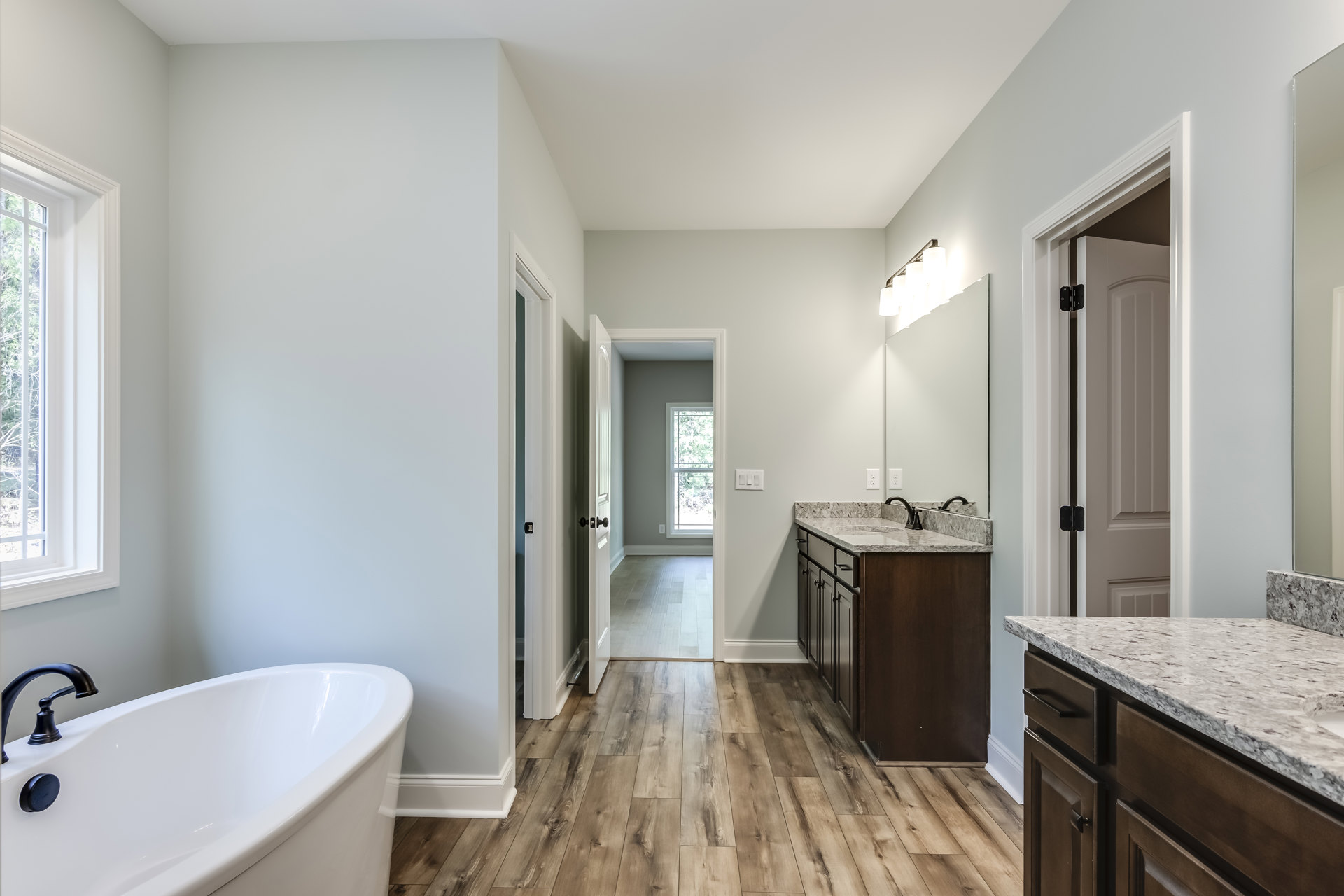 White freestanding bathtub with matte black shower head, large window overlooking trees, white countertop with integrated sink and black lamp, light tile flooring, minimalist
