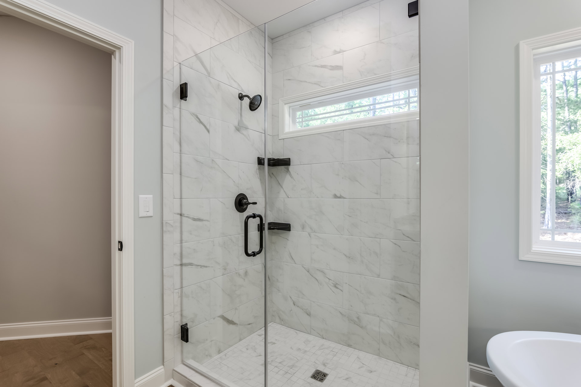Glass shower enclosure with tiled walls, large window overlooking trees, white toilet, chrome faucet, and black shower handle