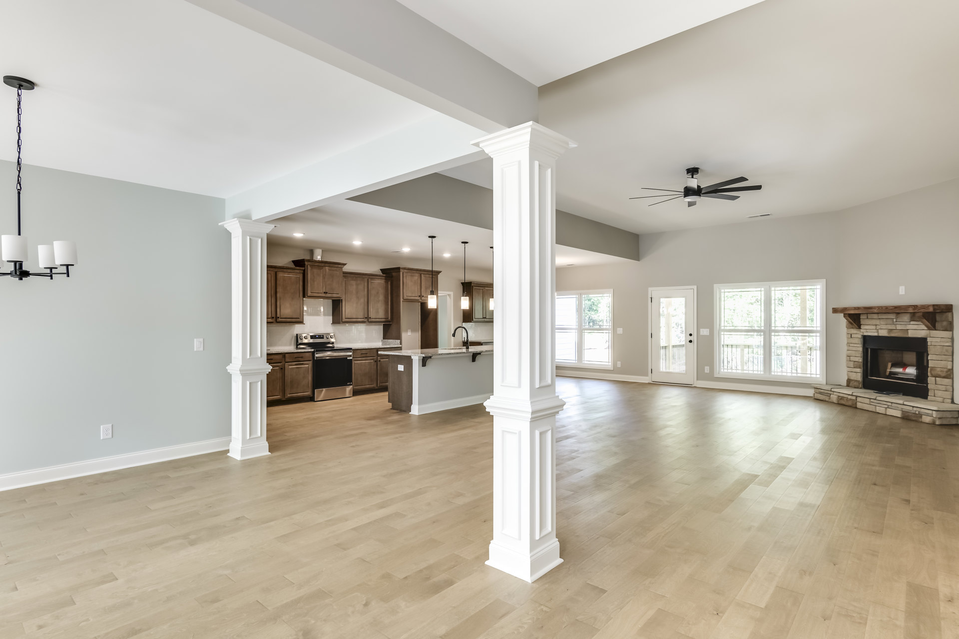 Spacious open floor plan featuring wood flooring, white columns, kitchen with glass door and silver hardware, dining area, fireplace with wood mantel, large window overlooking