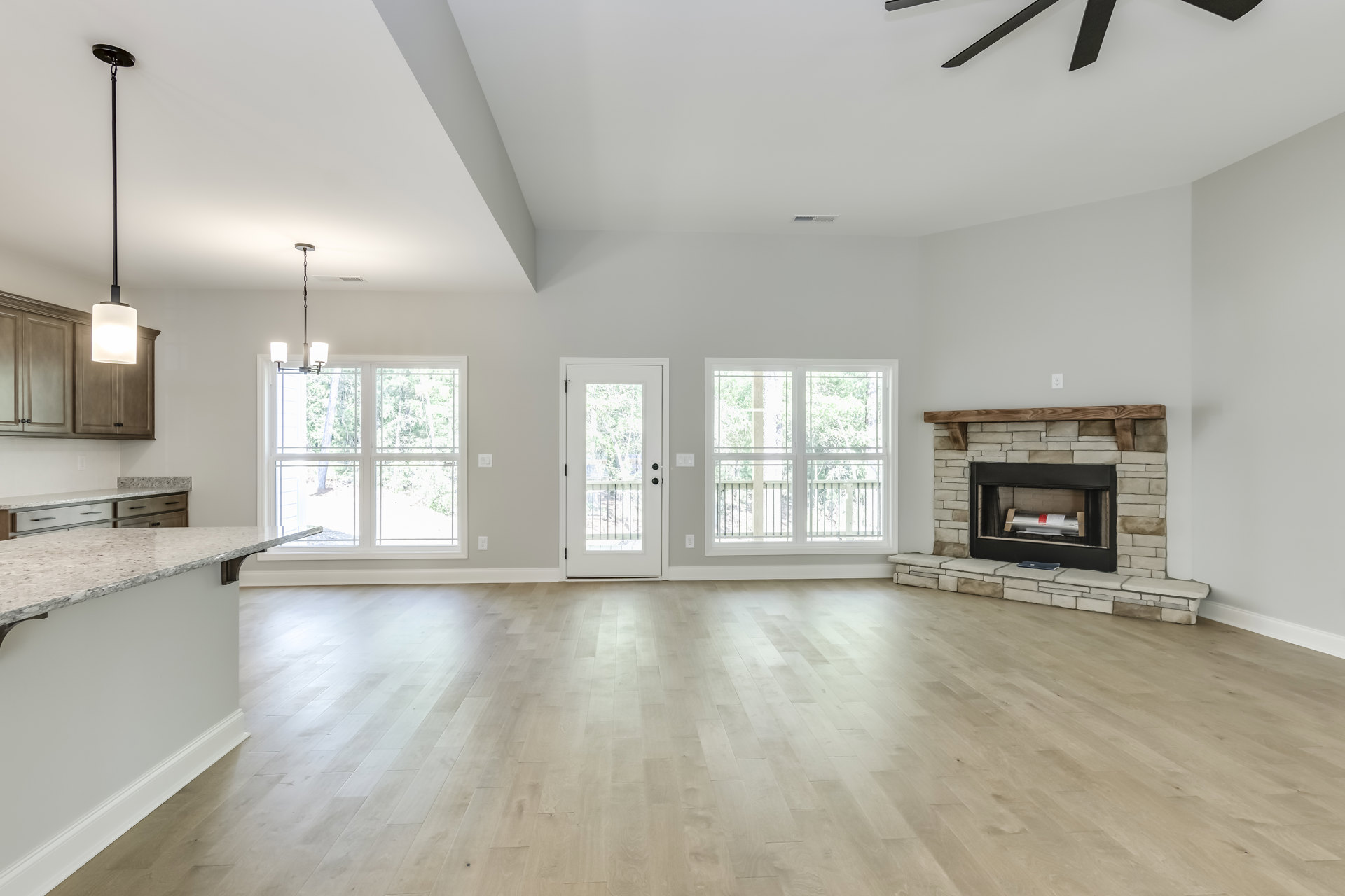 Living room with wood flooring, plaster walls, ceiling fan, built-in fireplace with silver log holder, glass-panel door, double doors, large window overlooking deck and trees.