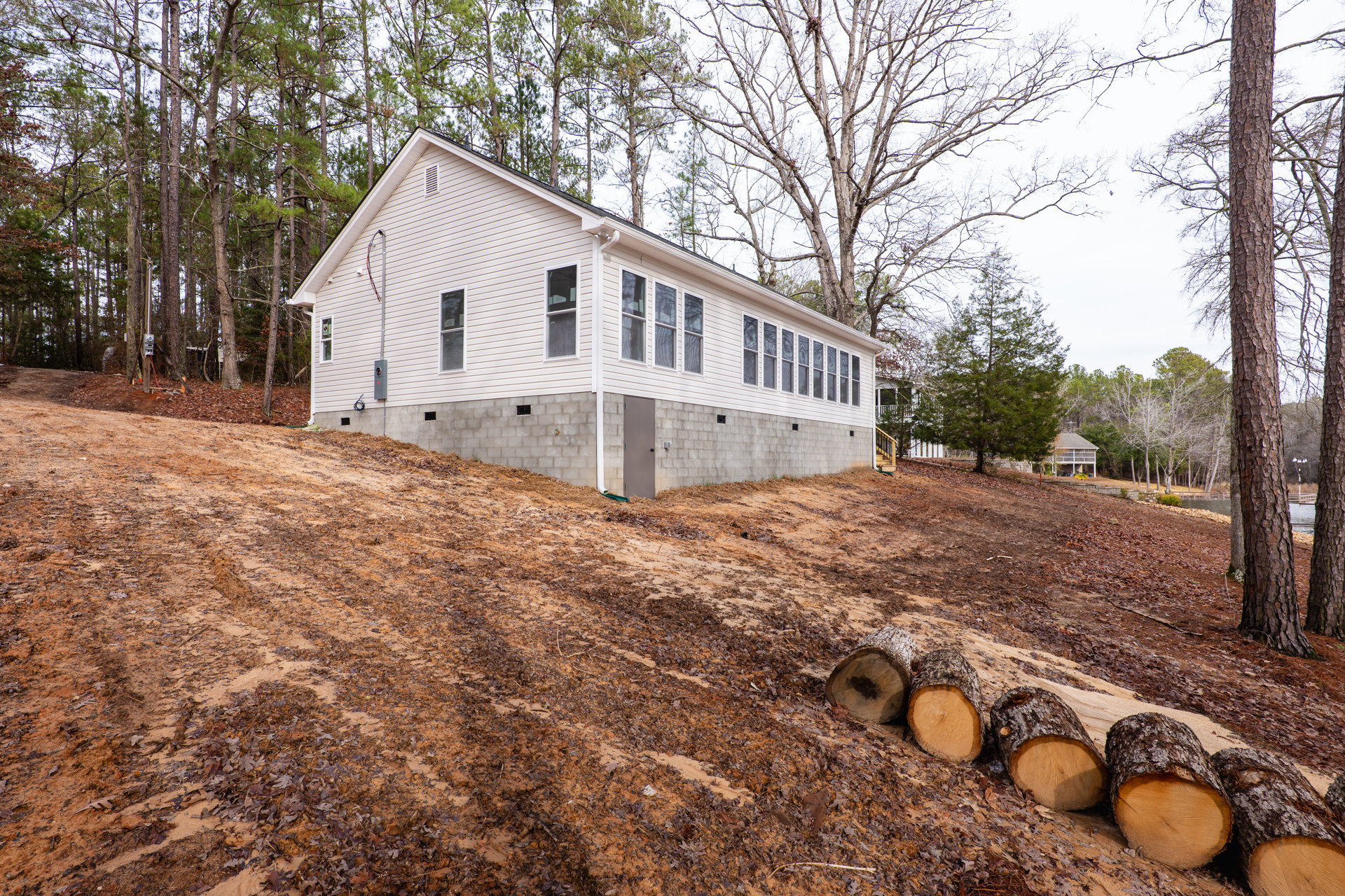 Two-story house with concrete block foundation, white siding, and windows, surrounded by trees; pile of cut logs resting on dirt and leaves in the front yard.