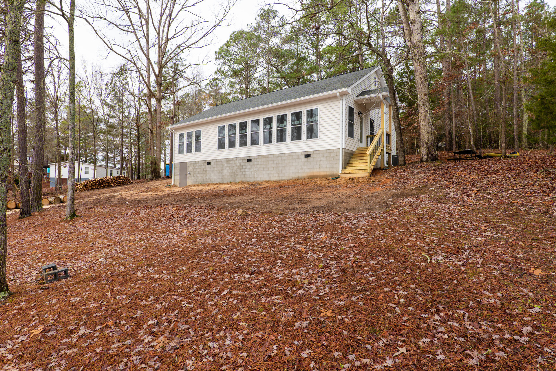 Modern house with large windows, wooden exterior staircase, surrounded by autumn leaves and dense forest, pile of firewood stacked beside concrete foundation