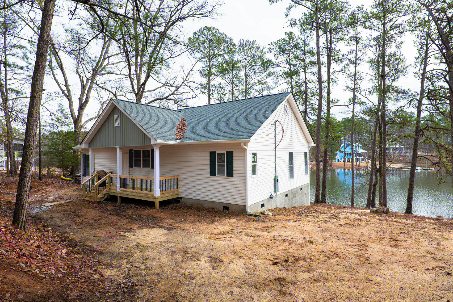 Two-story house with covered porch, black metal railing, wooden deck, large windows, tree with branching trunk in foreground, lake visible in background, roof scattered with fallen